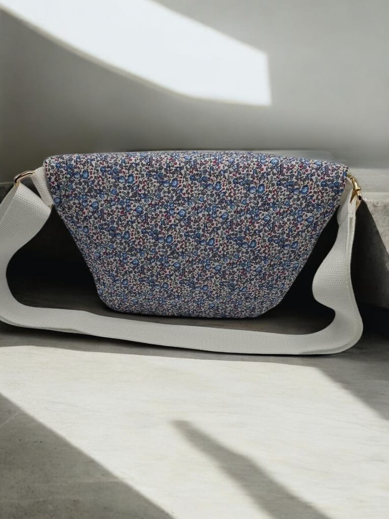Sac banane fleuri Liberty rouge avec bride blanche et fermeture dorée, posé sur une surface lumineuse.