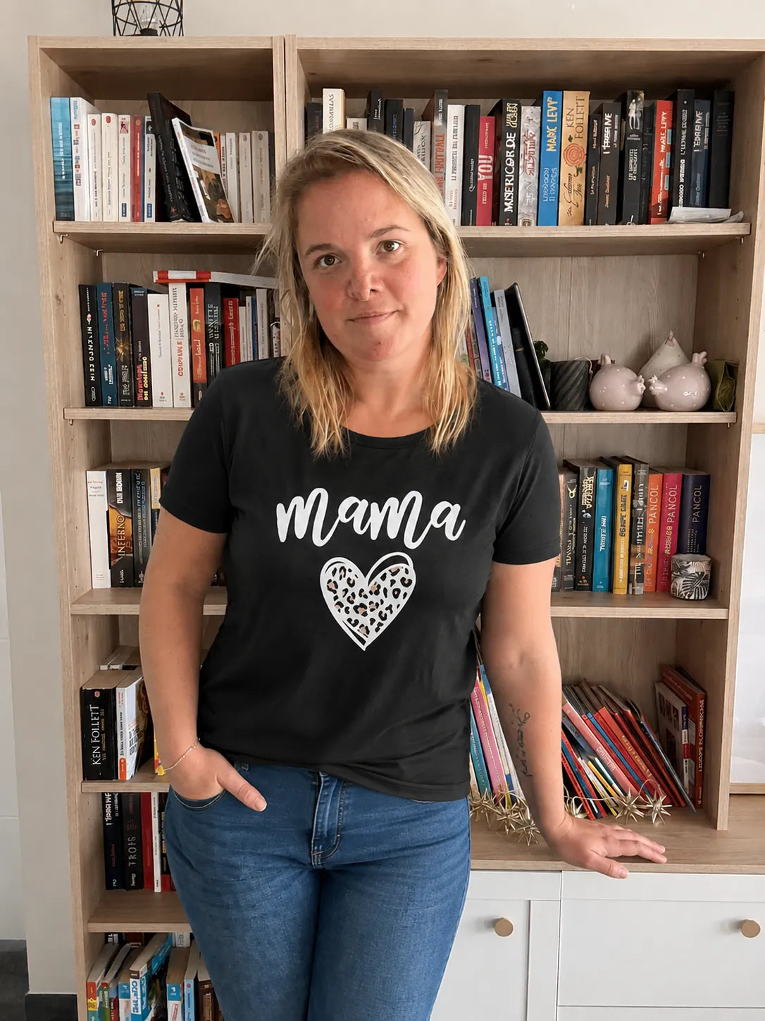 T-shirt noir femme avec le mot « mama » et cœur léopard, portée par une femme devant une bibliothèque.