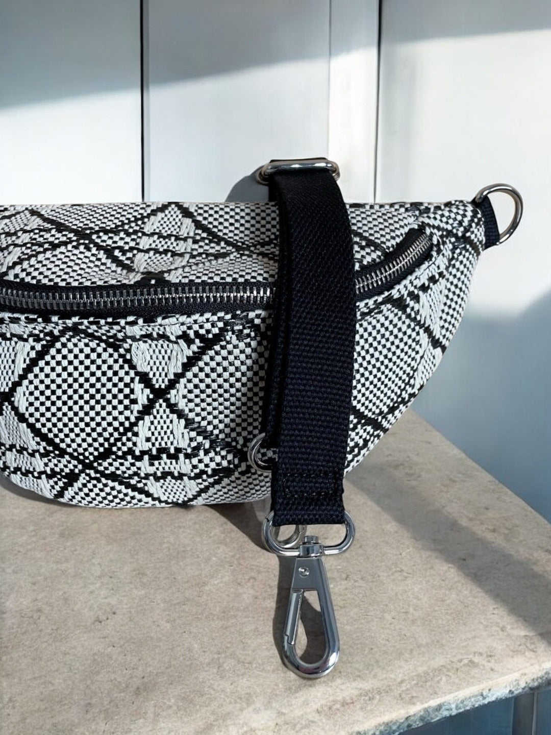 Sac banane effet tissu tressé noir et blanc avec bandoulière noire et fermeture éclair, posé sur une surface en marbre