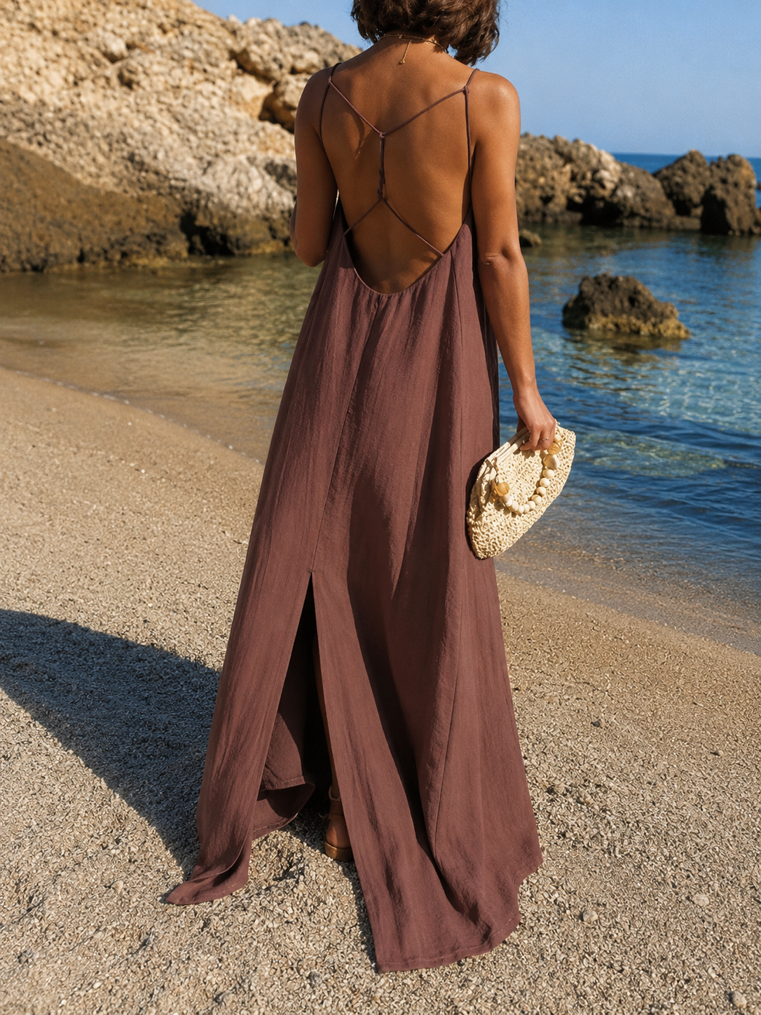 Robe été ample échancrée chocolat, dos nu, portée sur la plage avec des rochers et la mer en arrière-plan.