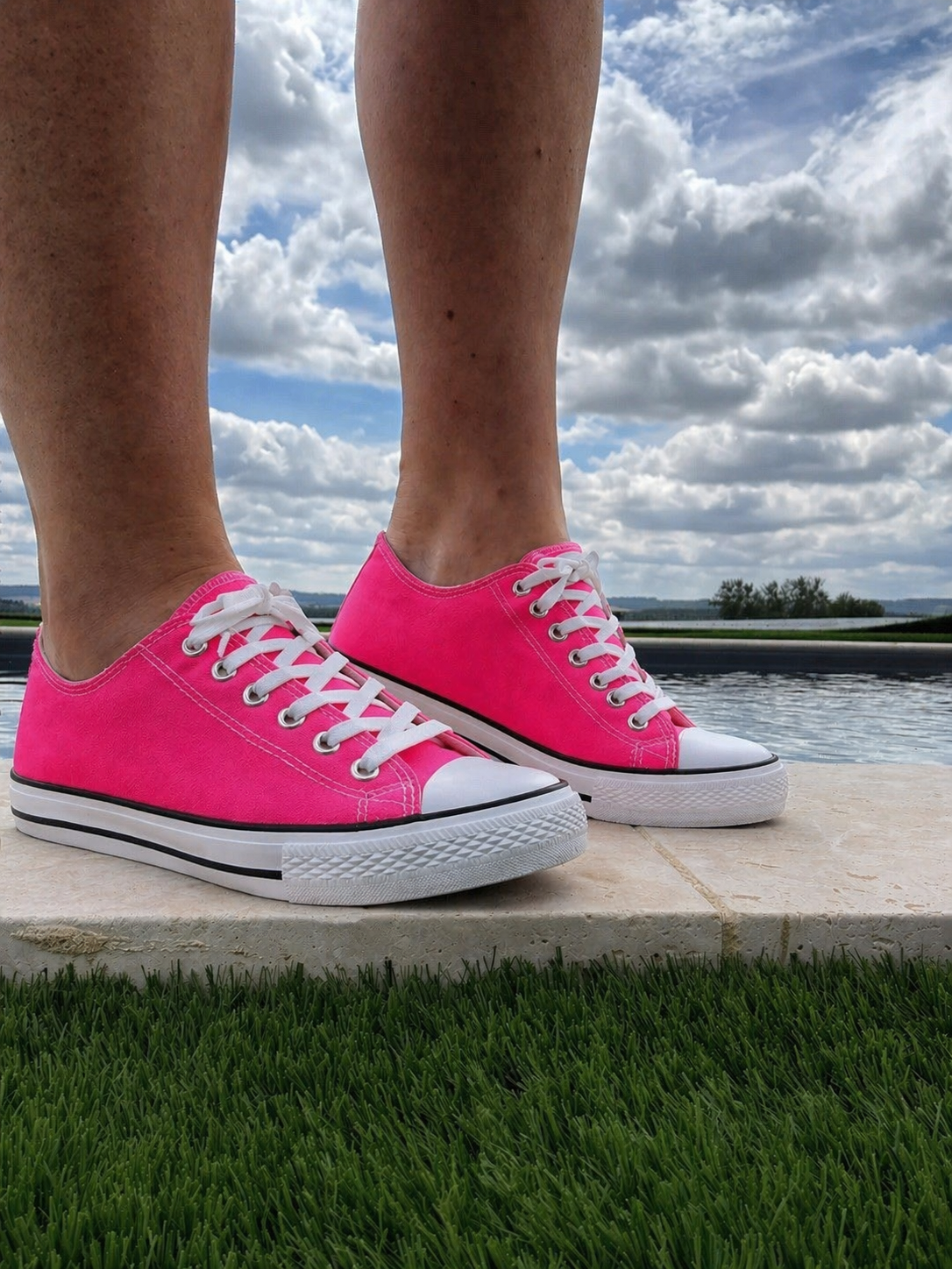 Paires de baskets roses fuchsia Converse-style posées sur une bordure en pierre près d’un plan d’eau, ciel nuageux en arrière-plan.