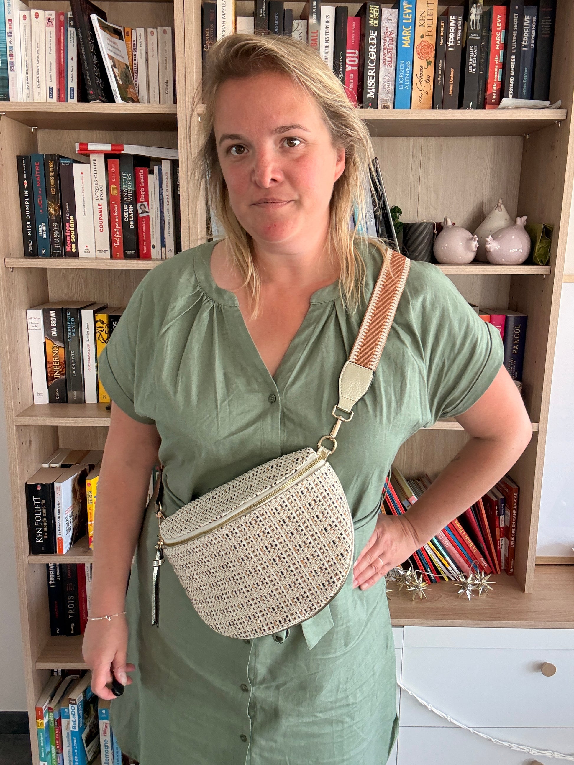 Sac banane en tissu au motif bohème, noir, beige et camel, porté en bandoulière par une femme devant une étagère remplie de livres.