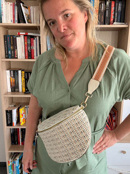 Sac banane tissé bohème portée en bandoulière, couleur naturel avec détails beige et camel, devant bibliothèque.