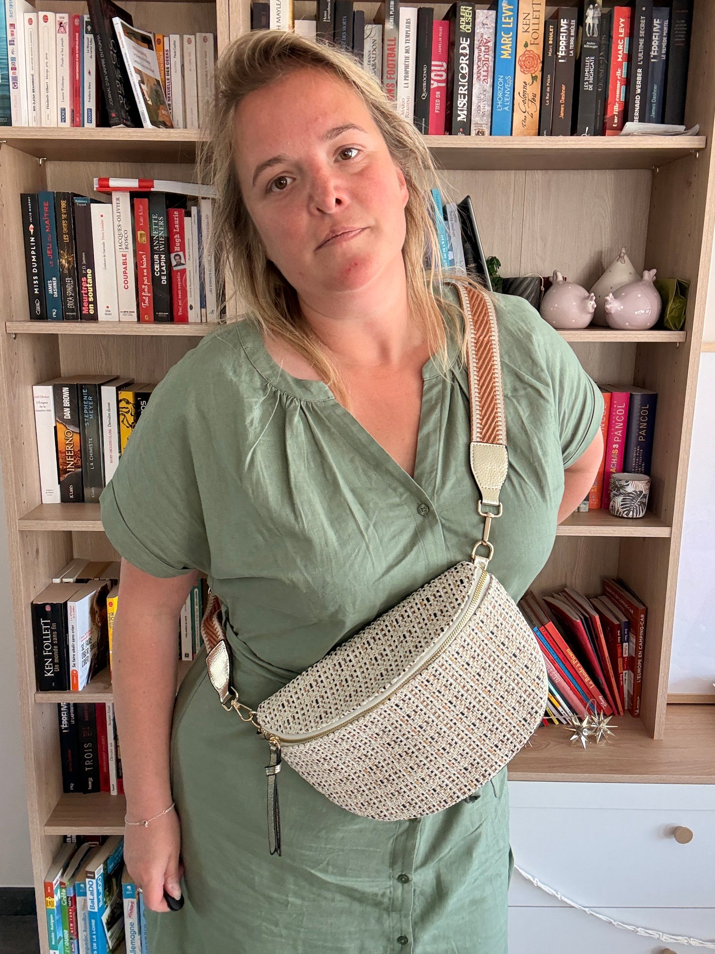 Homme ou femme portant sac banane tissé kaki/beige avec bandoulière motif bohème, devant étagères de livres.
