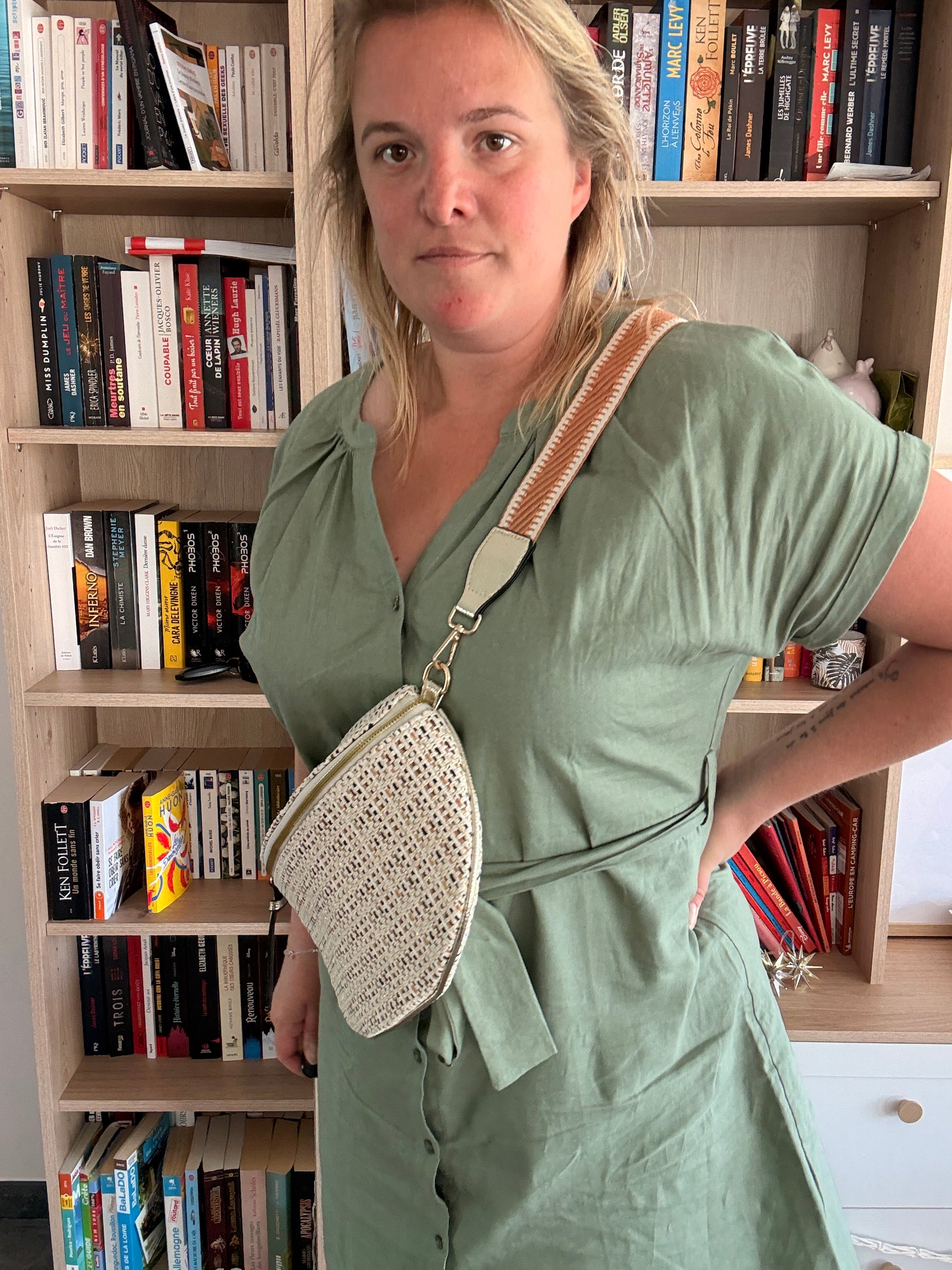Sac banane tissée noir, beige et camel porté en bandoulière par une femme devant une bibliothèque.