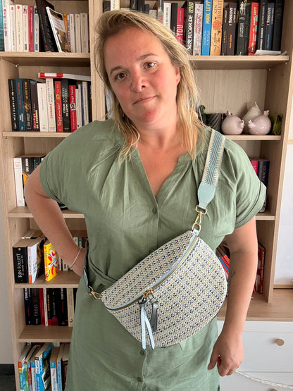 Sac banane en tissu à motif texturé couleur blanc cassé avec touches bleu clair, porté crossbody sur fond bibliothèque.