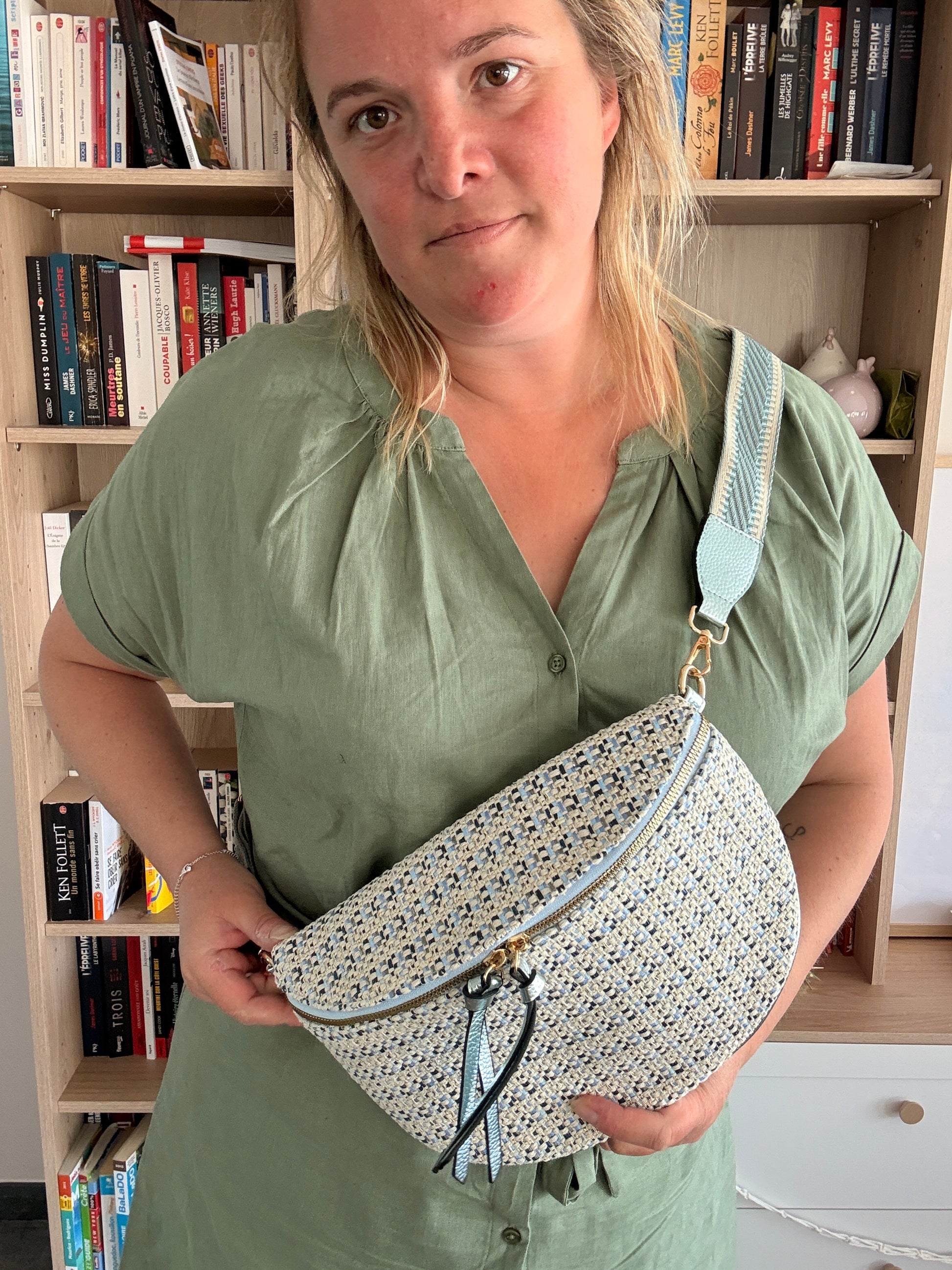 Sac banane en tissu à motif texturé noir, bleu et beige porté en bandoulière par une personne devant une bibliothèque.