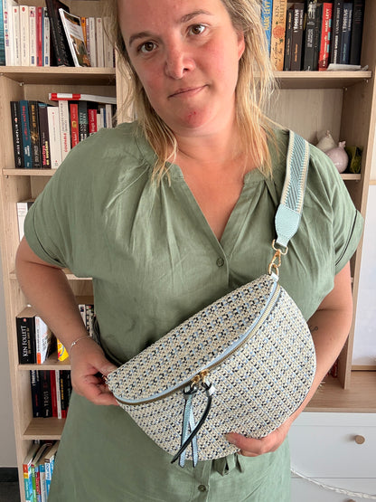 Sac banane en tissu à motif texturé noir, bleu et beige porté en bandoulière par une personne devant une bibliothèque.
