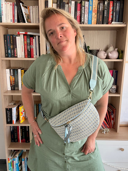 Sac banane en tissu à motif texturé noir, bleu et beige porté en bandoulière sur une femme devant une étagère remplie de livres.