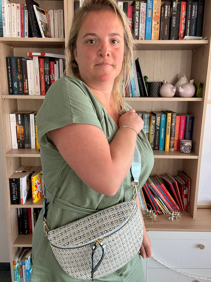 Sac banane en tissu à motif texturé noir, bleu et beige porté en bandoulière par une femme devant une bibliothèque.