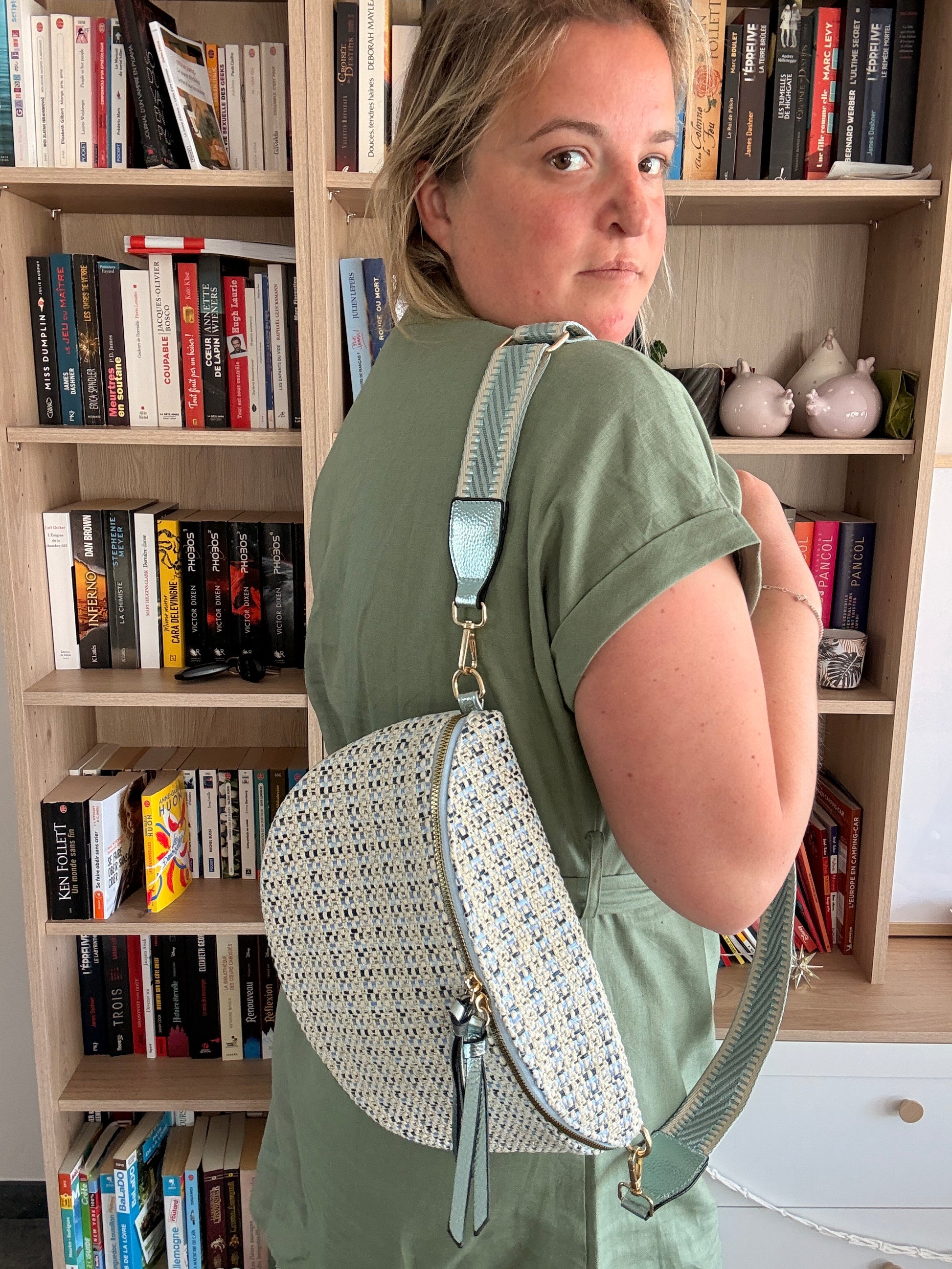 Sac banane en tissu à motif texturé de couleurs noir, bleu et beige porté en bandoulière sur l’épaule d’une personne, fond bibliothèque.