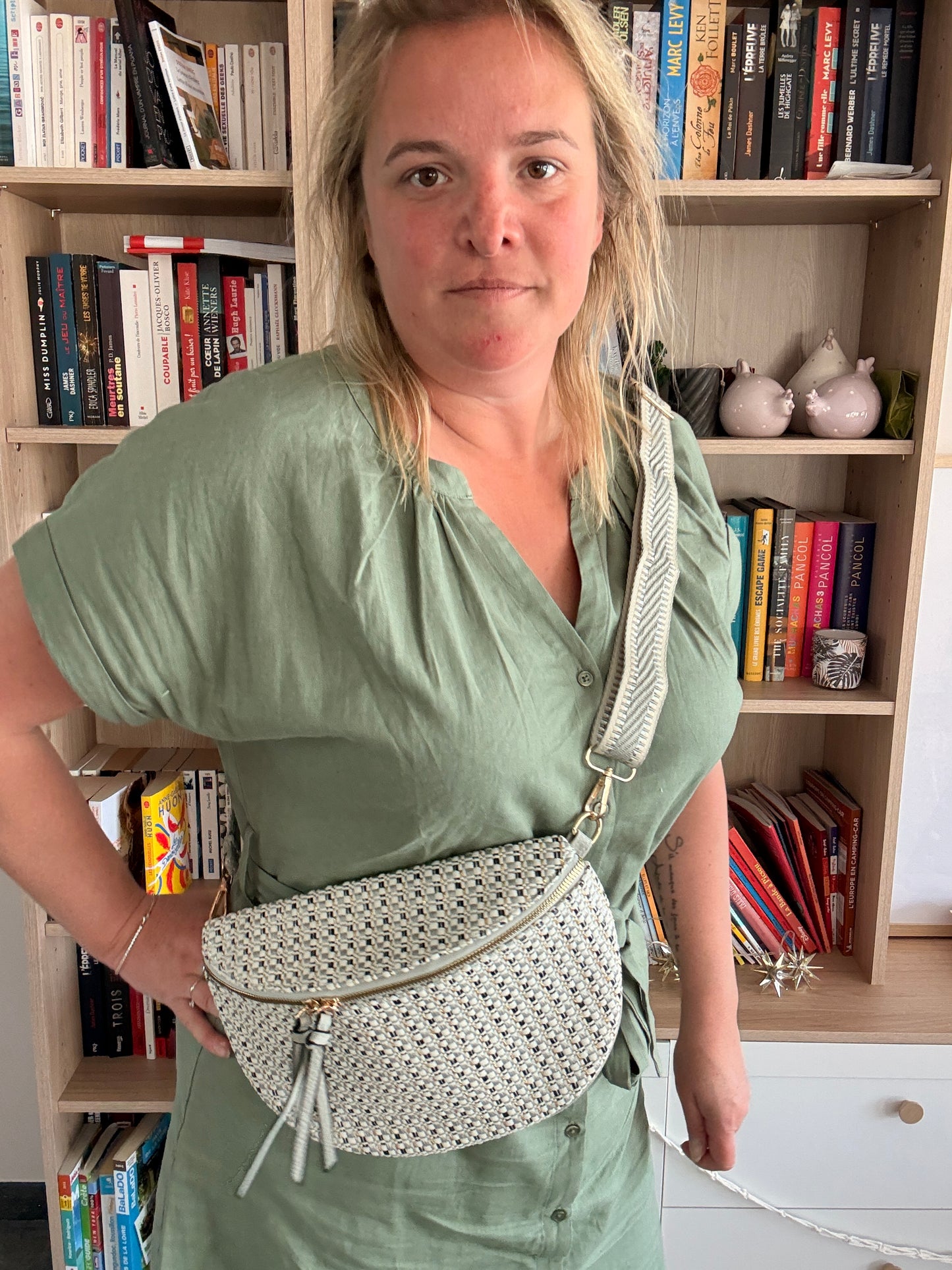 Sac banane en tissu texturé avec motif clair, portée en bandoulière par une femme devant une étagère de livres.