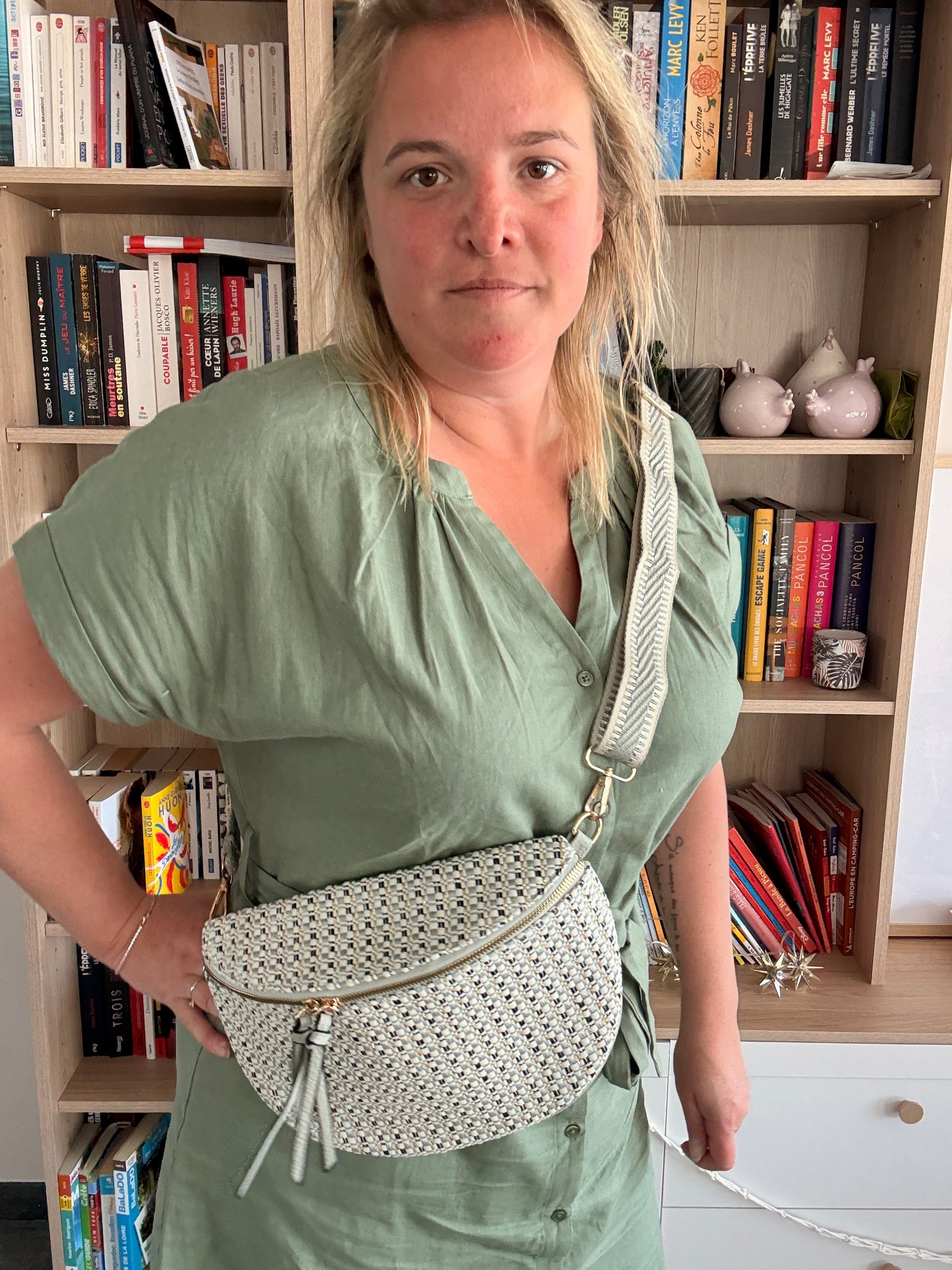 Sac banane en tissu texturé avec motif clair, portée en bandoulière par une femme devant une étagère de livres.