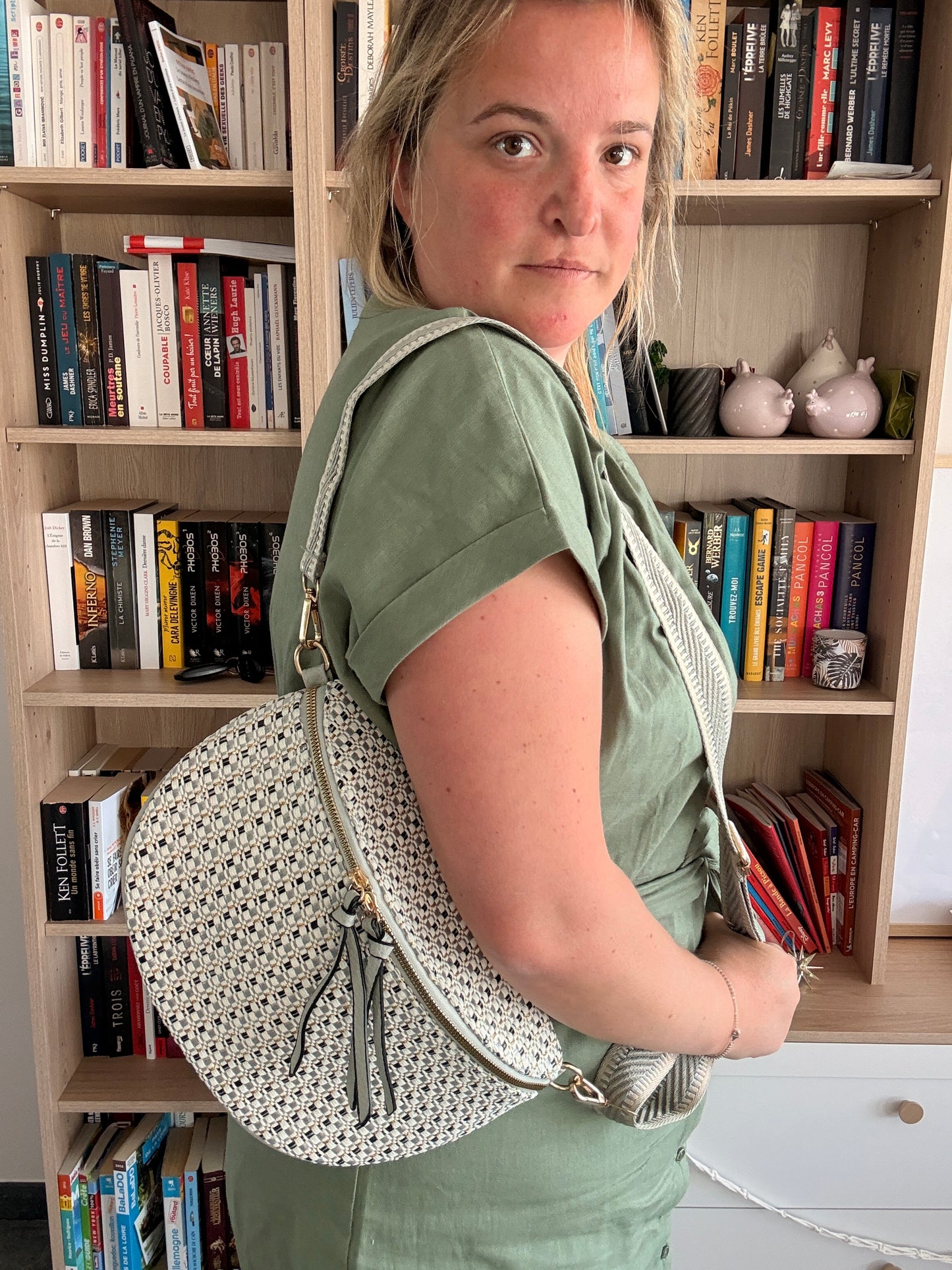 Sac banane en tissu texturé à motifs blanc, noir et bleuté porté sur l’épaule d’une femme, en arrière-plan étagères de livres.