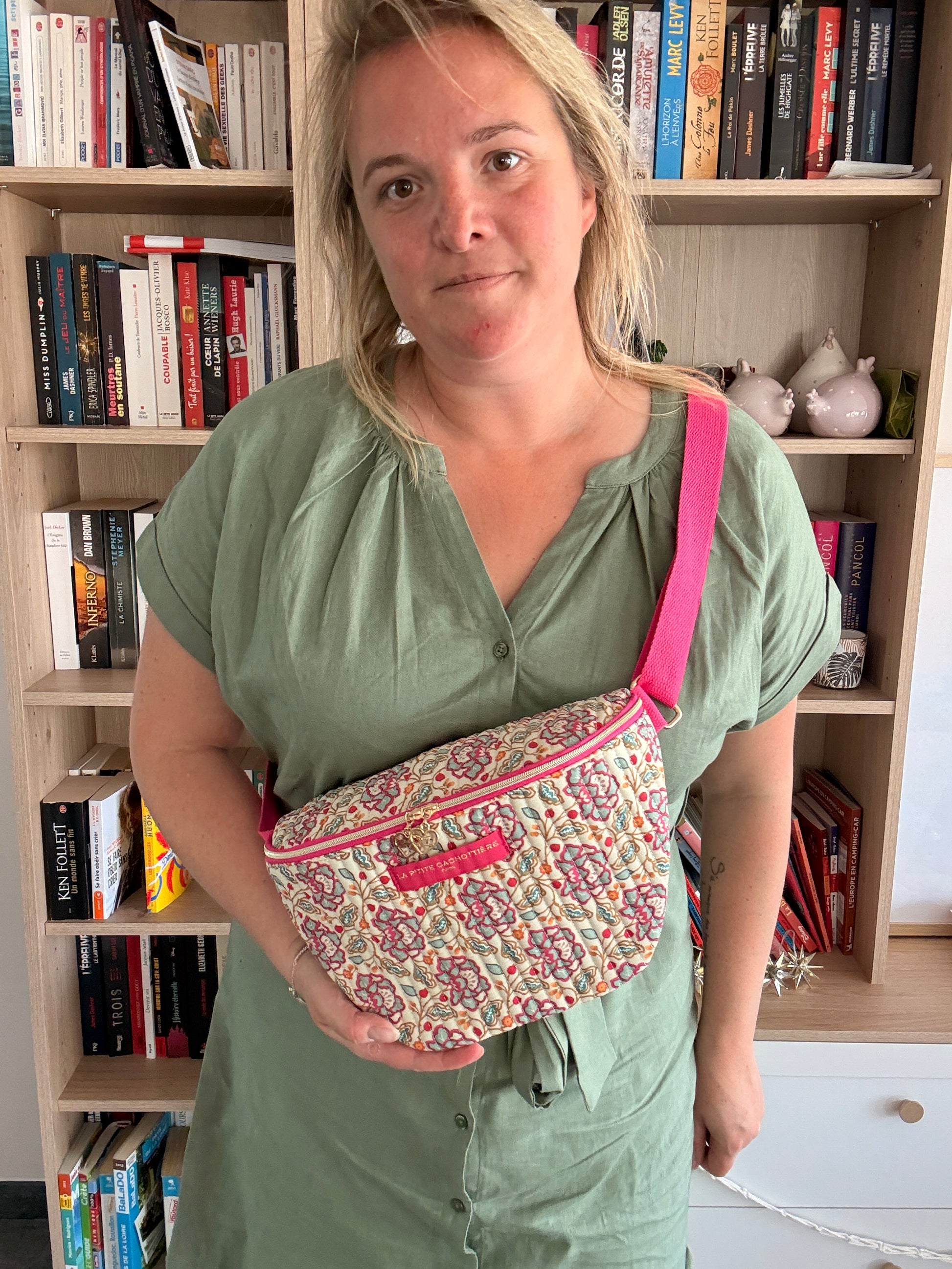Sac banane Liberty fleuri rose portée en diagonale par une femme, fond bibliothèque. Couleur beige et dorures visibles sur le motif. Présente une sangle rose et un design floral.