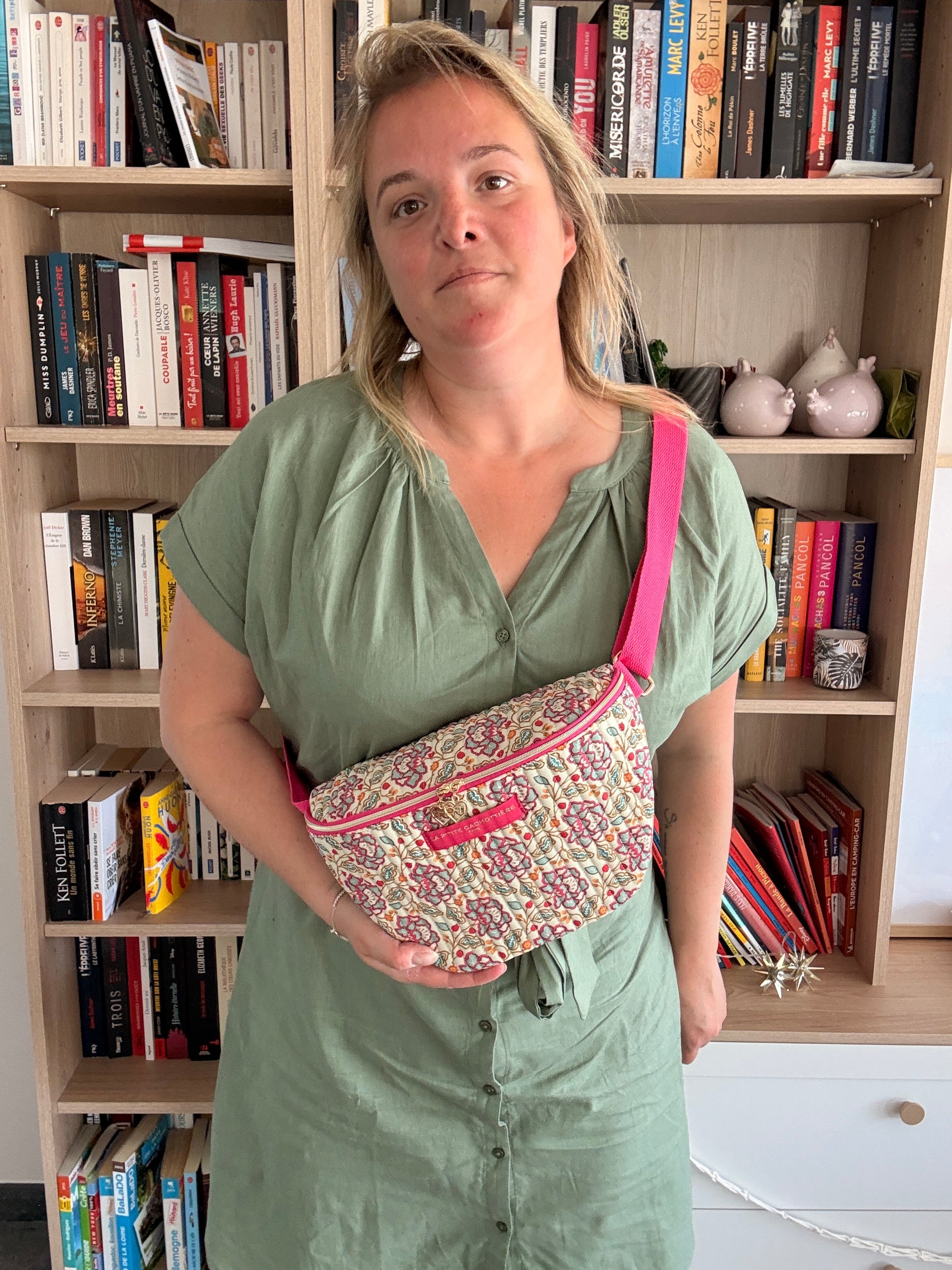 Sac banane Liberty fleuri rose porté diagonale sur l’épaule d’une femme, robe vert olive, bibliothèque en arrière-plan.
