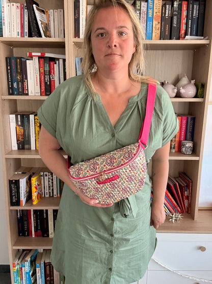 Sac banane Liberty fleuri rose porté en bandoulière par une femme, avec une ceinture beige et dorures, devant une étagère de livres.