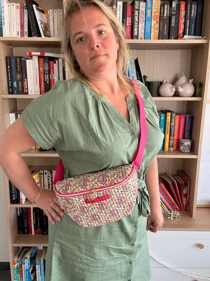 Sac banane Liberty fleuri rose porté en travers, sur un vêtement vert clair, avec une sangle rose et un fond bibliothèque.