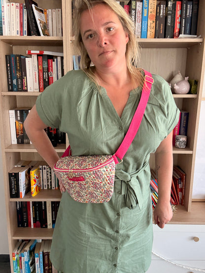 Sac banane Liberty fleuri rose porté en bandoulière sur l’épaule d’une femme près d’une bibliothèque, coton beige et finitions dorées et rose vif.
