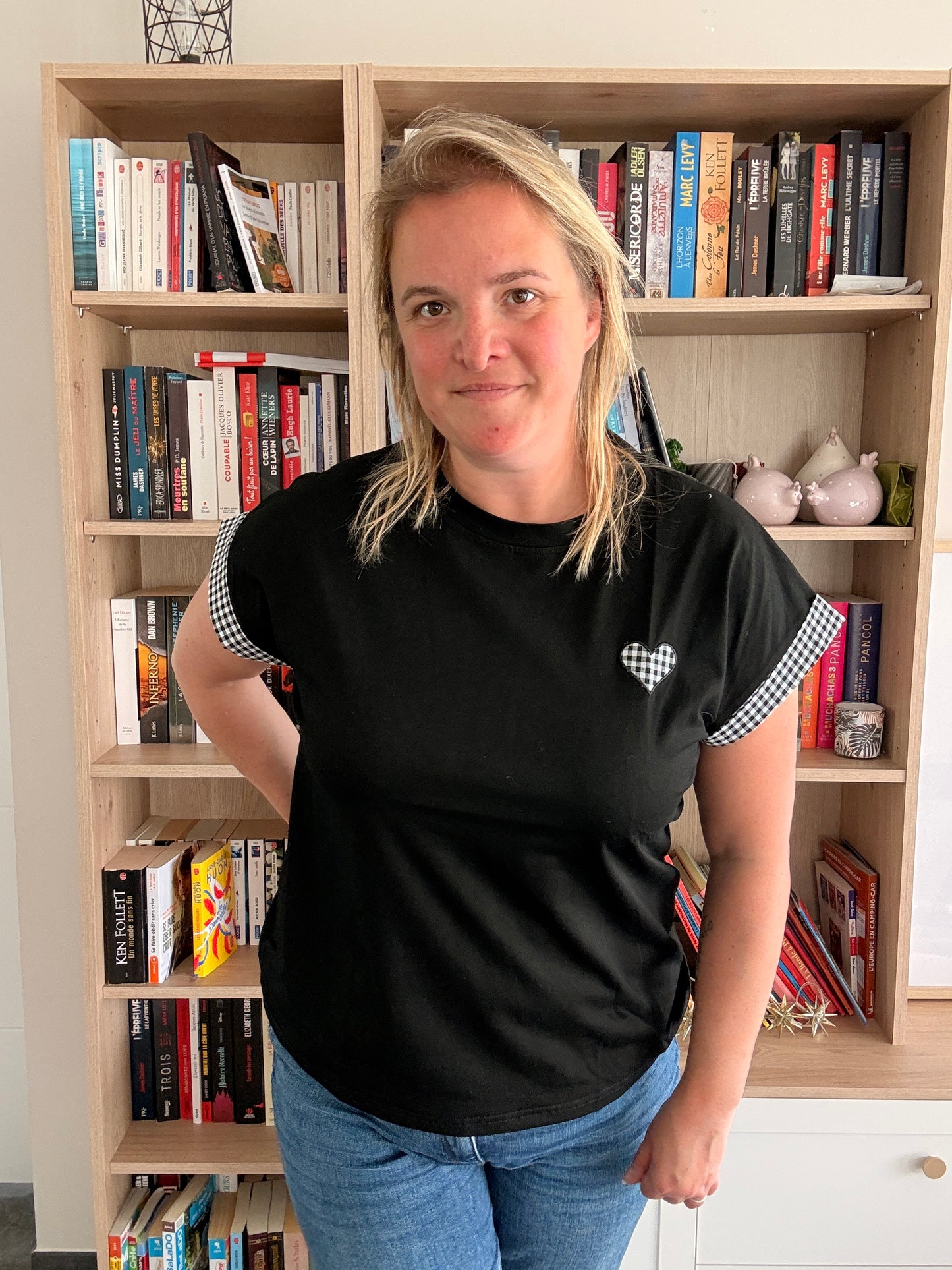 Femme portant un T-shirt noir oversize à détail Vichy noir et blanc sur le devant, en intérieur près d'une bibliothèque.