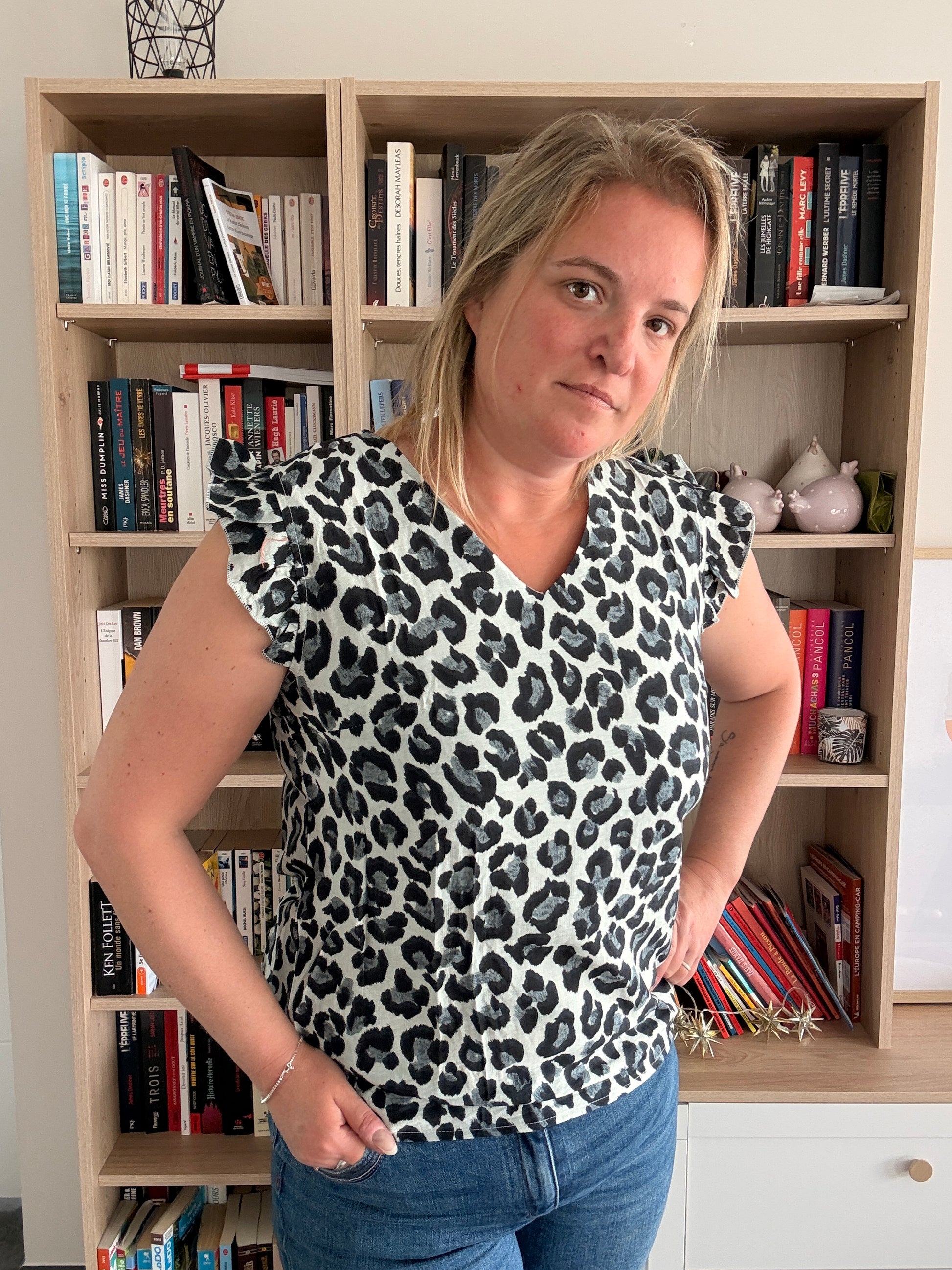 T-shirt femme sans manches à volants léopard noir et gris, porté par une femme debout dans une pièce avec bibliothèque.