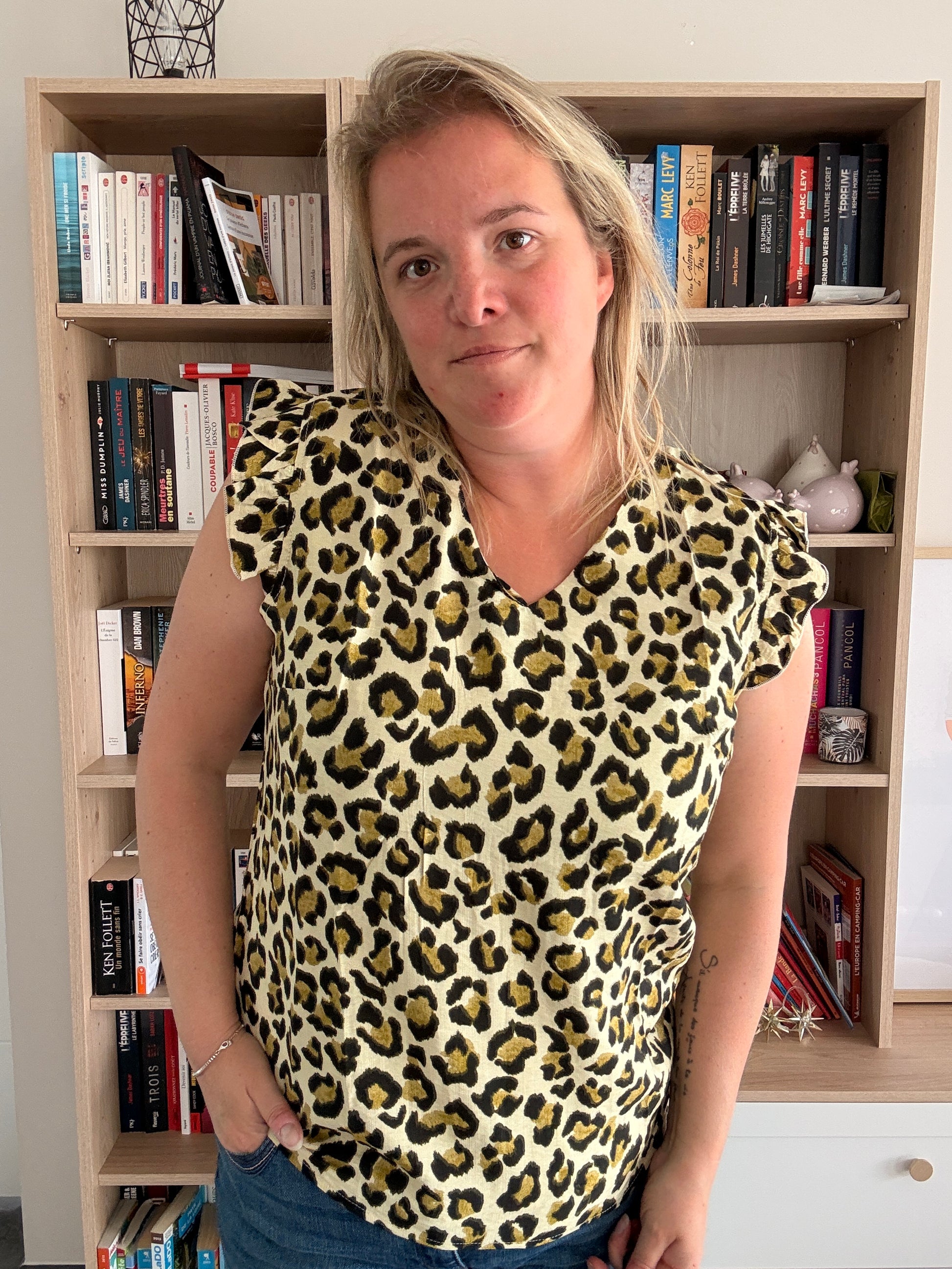 T-shirt femme sans manches à volants vert kaki effet léopard porté par une femme, debout devant une bibliothèque.