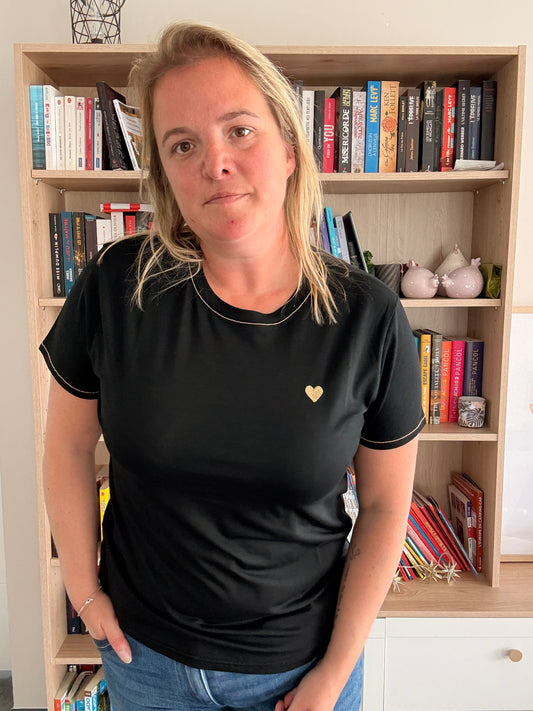 T-shirt noir avec petit cœur doré sur la poitrine porté par une femme debout devant une étagère remplie de livres.