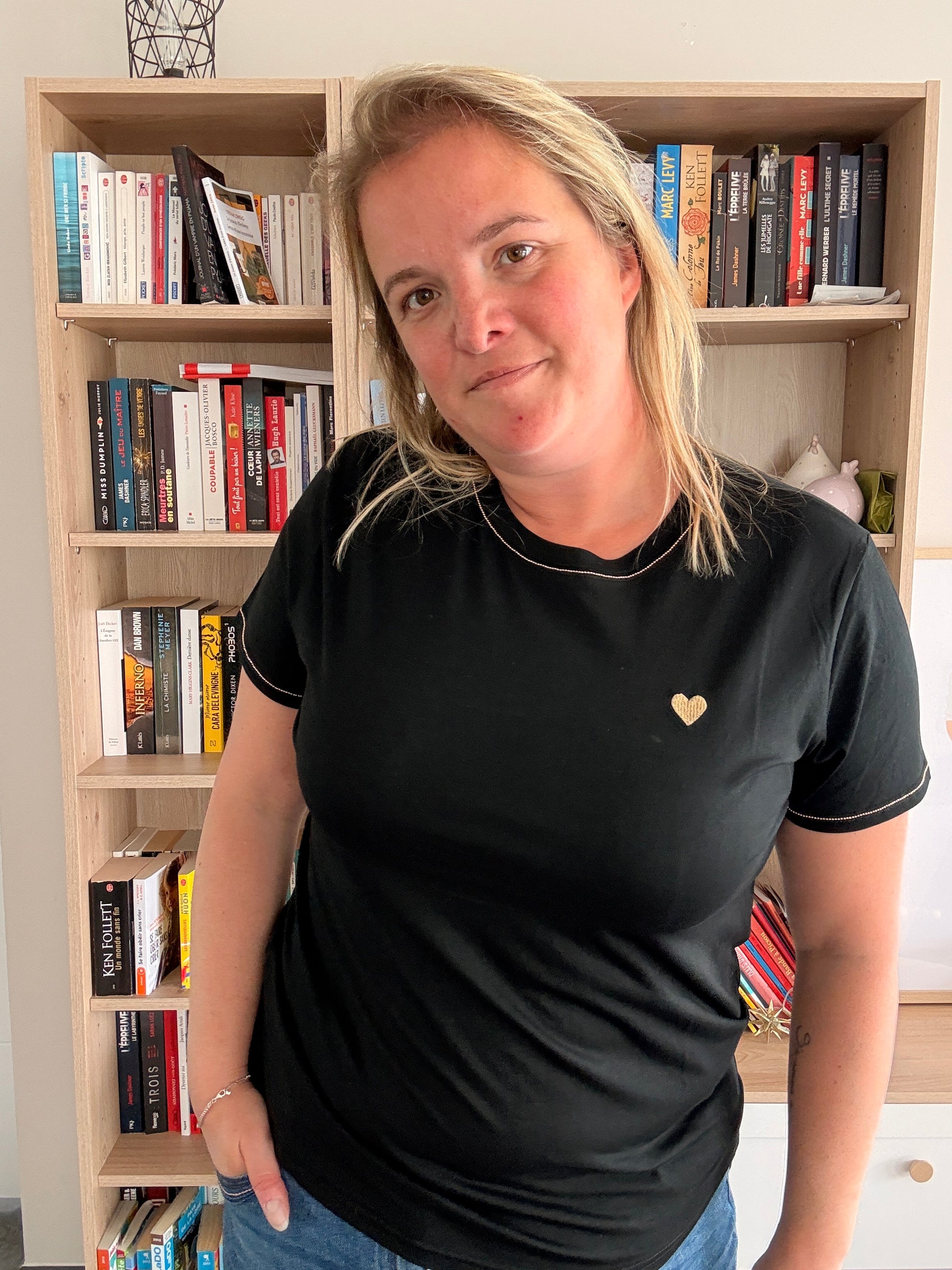 Homme ou femme portant T-shirt Noir Cœur Doré, debout devant une étagère de livres, sourire léger.