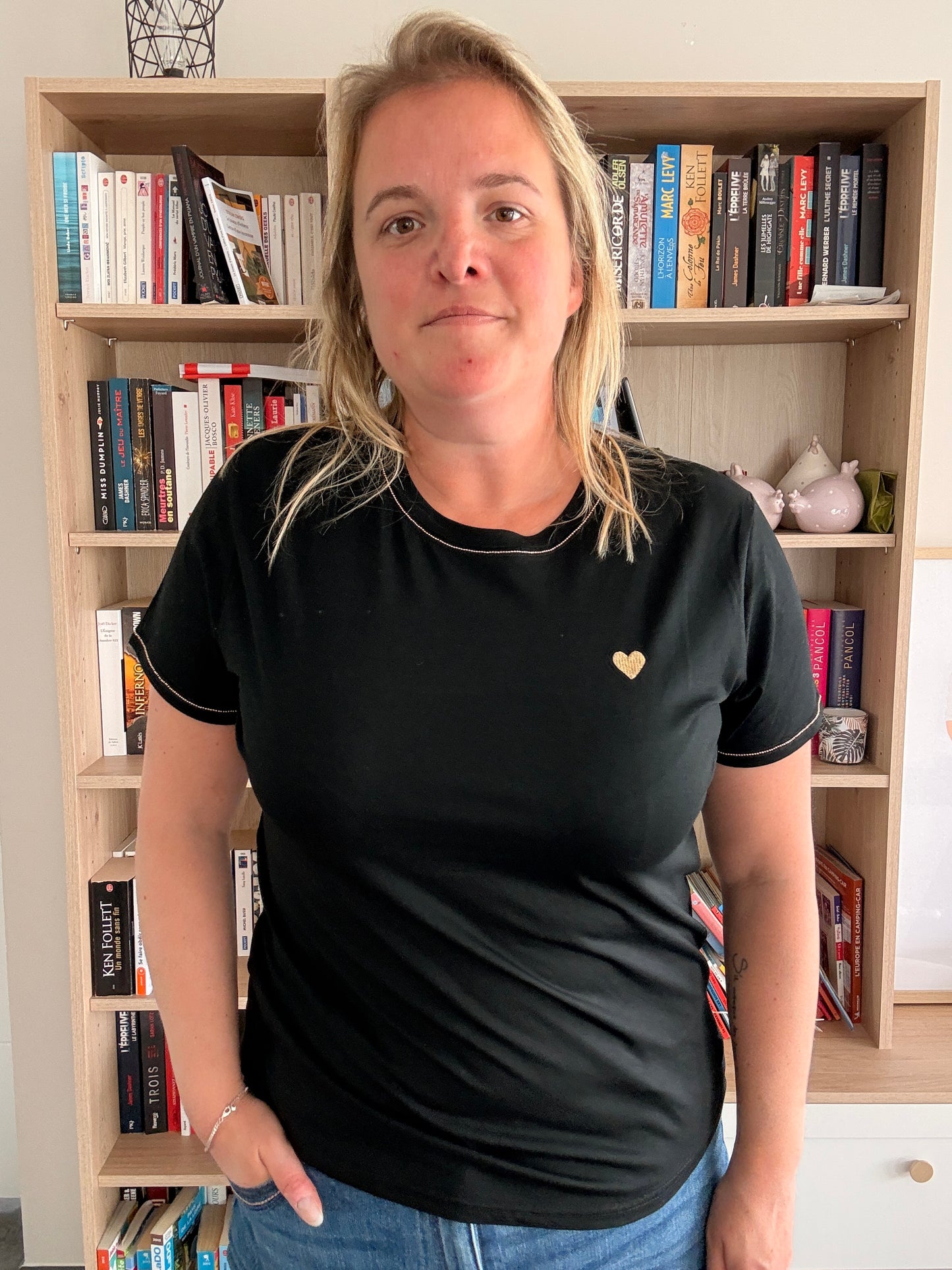 Homme ou femme portant un T-shirt noir avec cœur doré sur la poitrine, debout devant une bibliothèque.