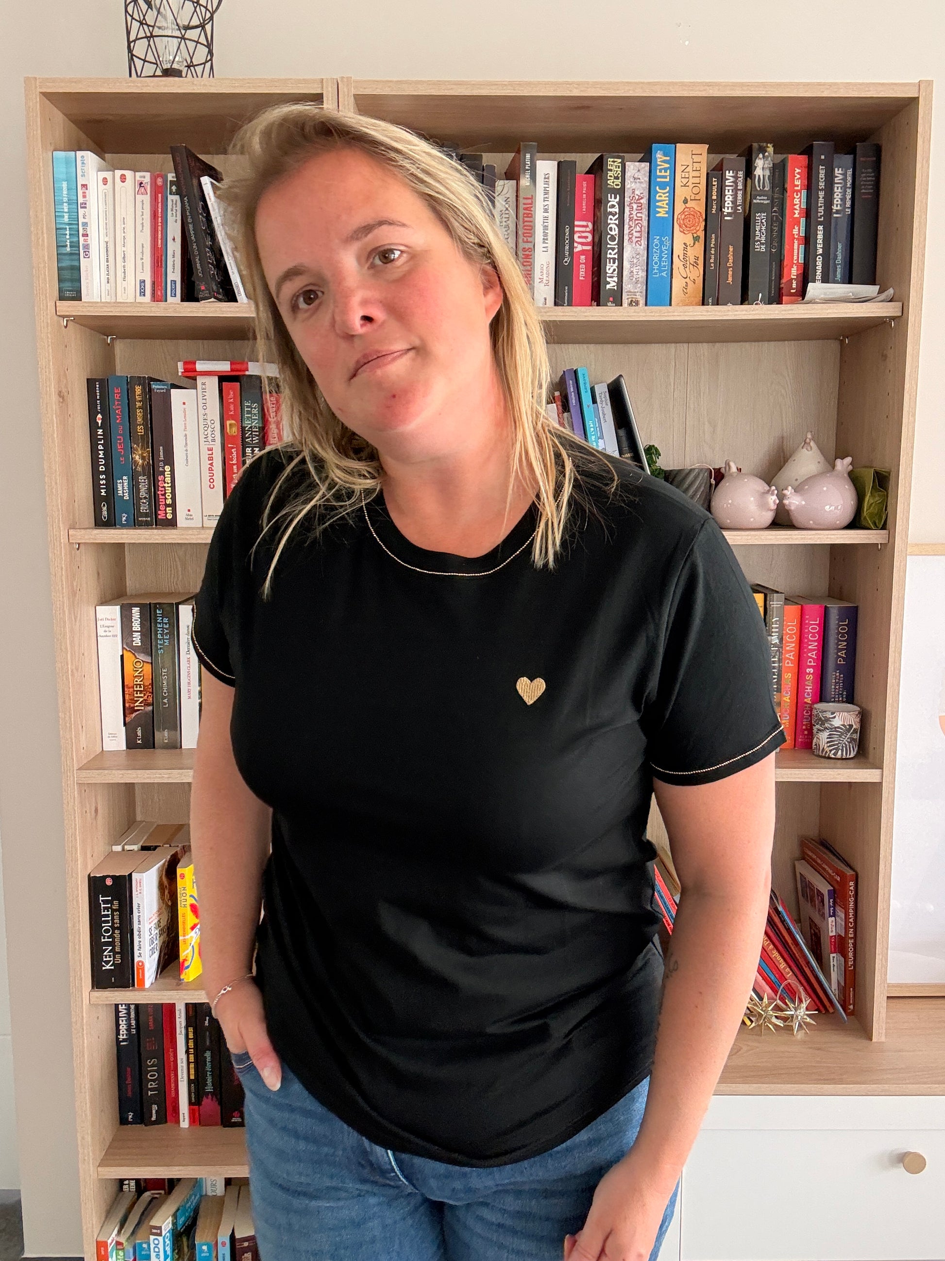 T-shirt noir avec cœur doré porté par une femme devant une bibliothèque.