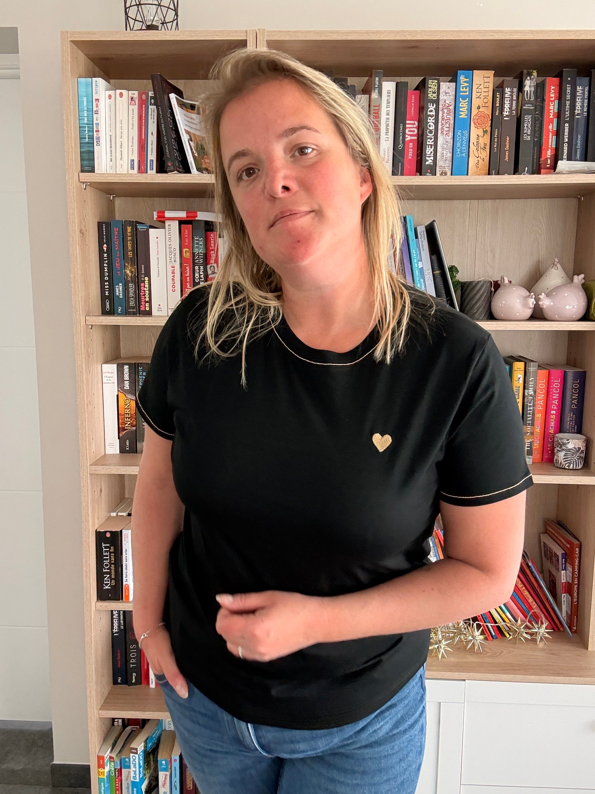 Homme ou femme debout porte un T-shirt noir avec cœur doré, er recliné, bibliothèque en arrière-plan.