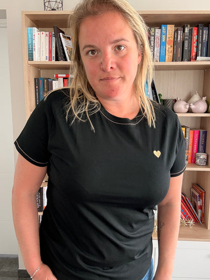 Homme ou femme portant un t-shirt noir T-Shirt Noir Cœur Doré, debout devant une bibliothèque, cœur doré sur le torse.