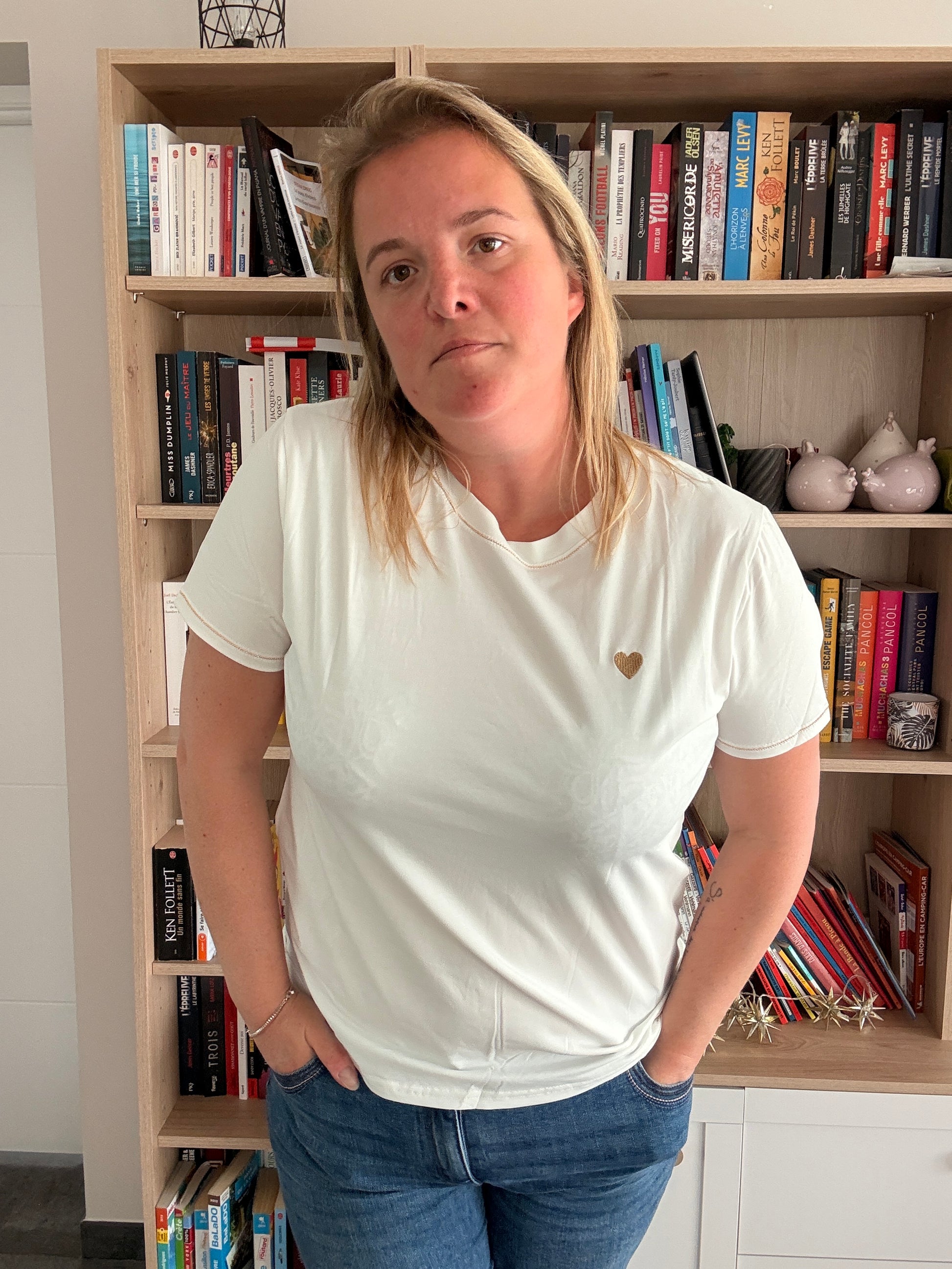 Homme ou femme portant un T-shirt blanc avec petit cœur doré sur la poitrine, debout devant une bibliothèque.