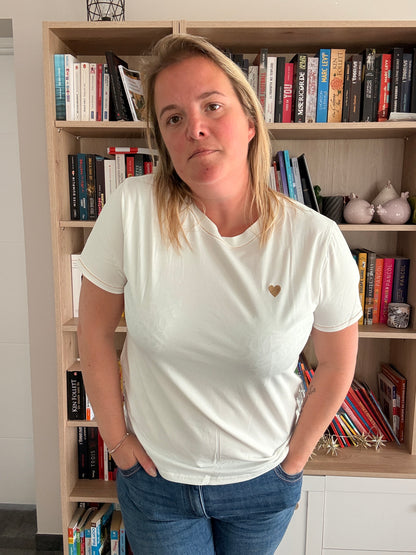 Homme ou femme portant un T-shirt blanc avec petit cœur doré sur la poitrine, debout devant une bibliothèque.