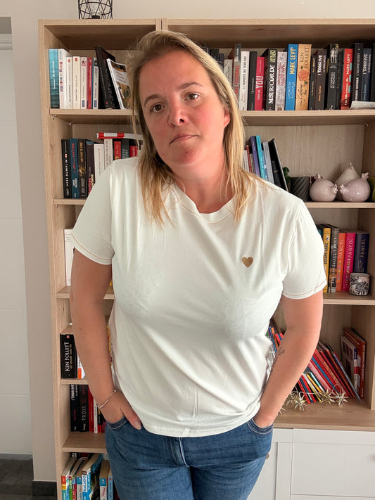 Homme ou femme portant un T-shirt blanc avec petit cœur doré sur la poitrine, debout devant une bibliothèque.