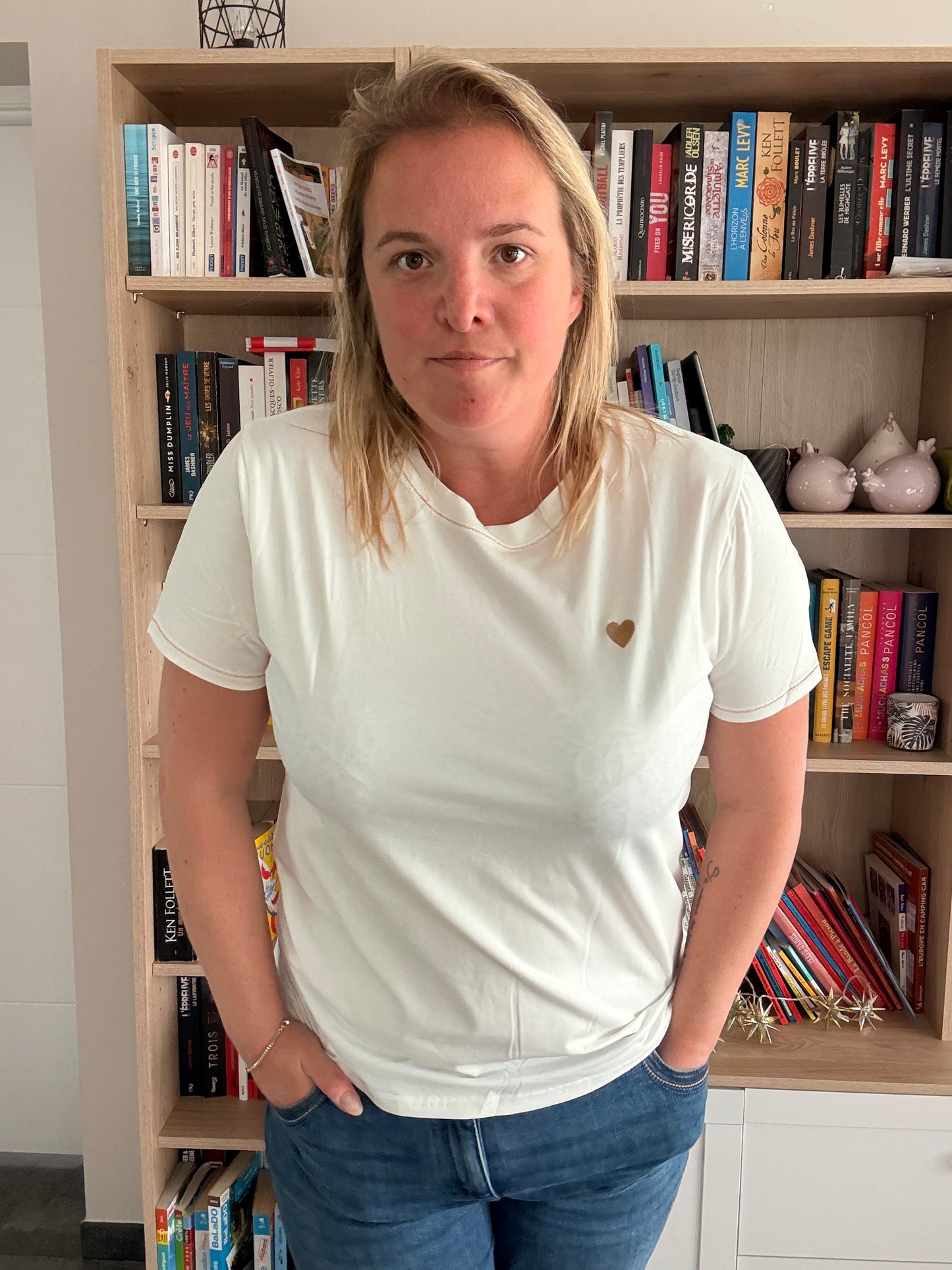 Homme ou femme portant T-shirt blanc avec petit cœur doré sur la poitrine, debout devant une étagère de livres.