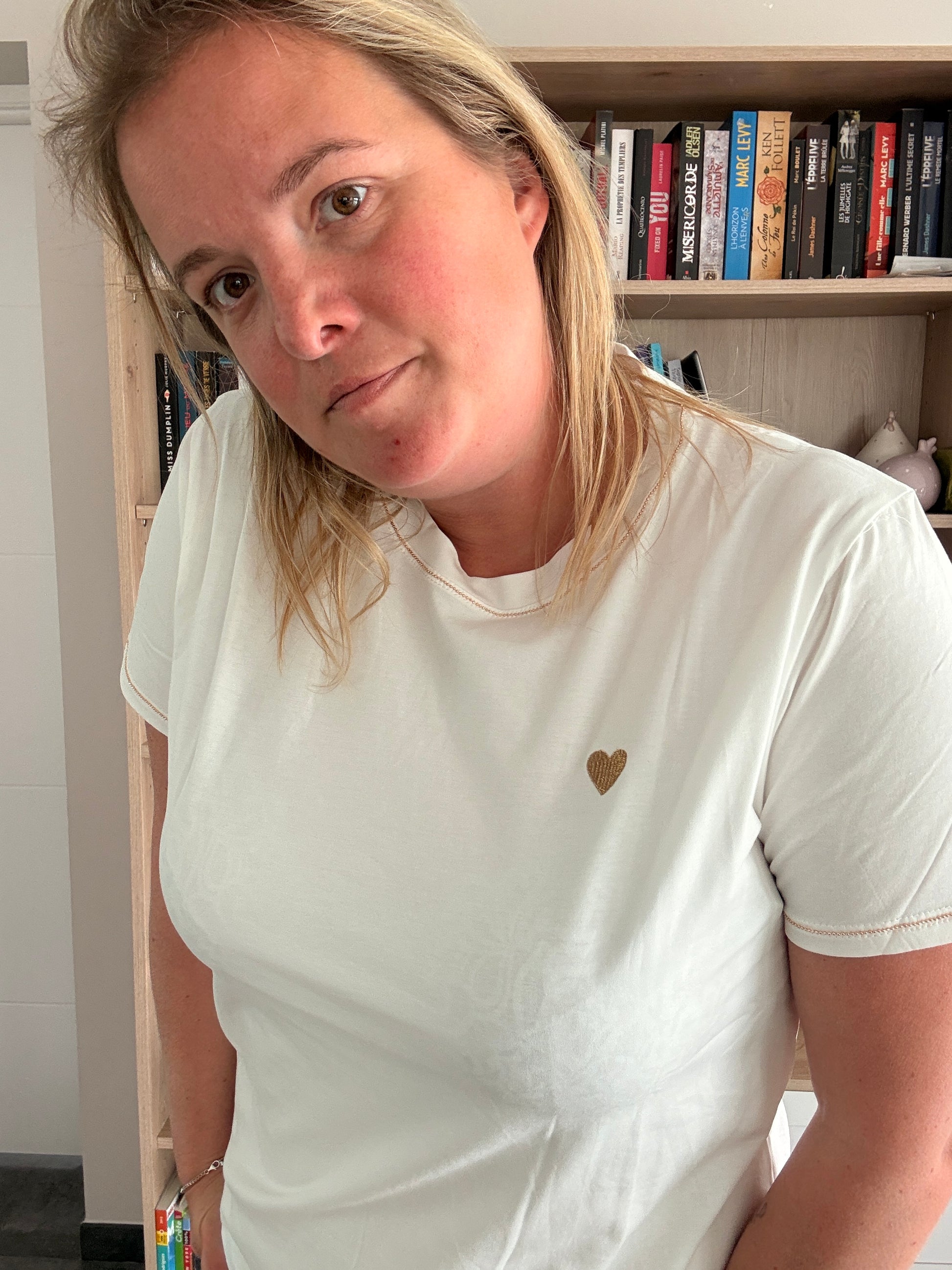 T-shirt blanc avec petit cœur doré sur la poitrine, porté par une femme dans une pièce avec étagères et livres en arrière-plan.