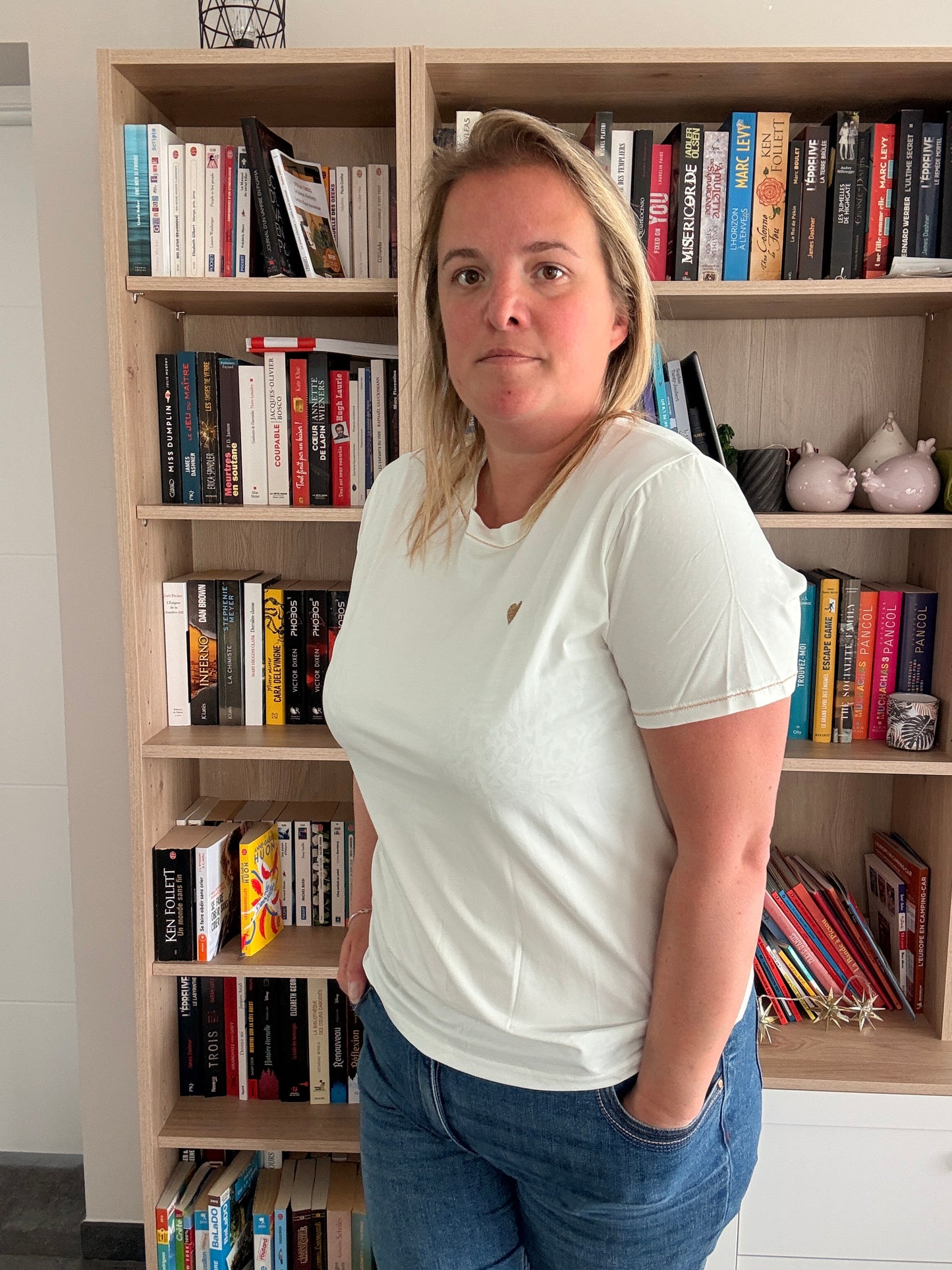 Femmes décontractée en T-shirt blanc Cœur Doré, debout dans une bibliothèque remplie de livres.