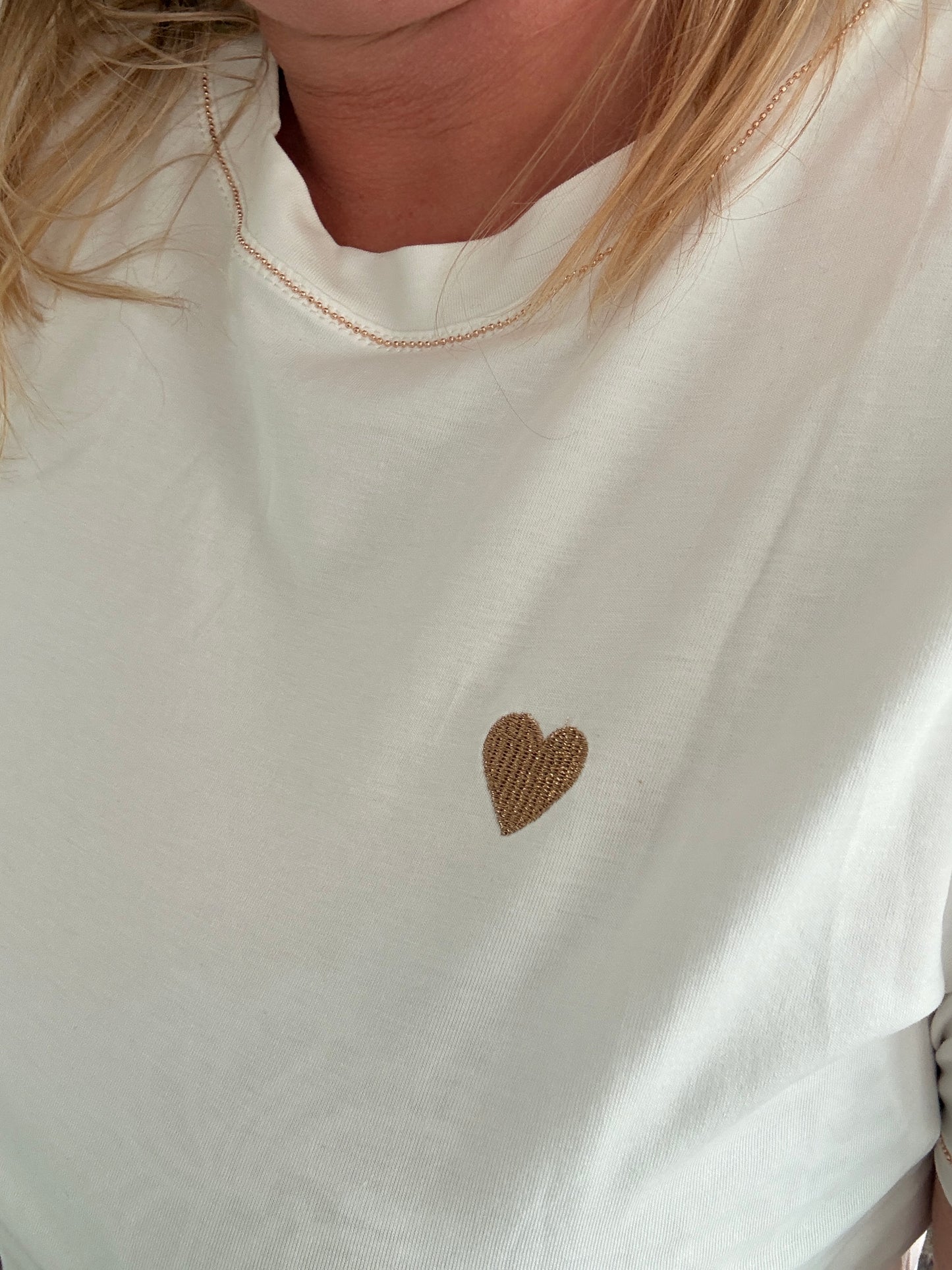 T-shirt blanc avec petit cœur doré sur la poitrine, porté par une personne.