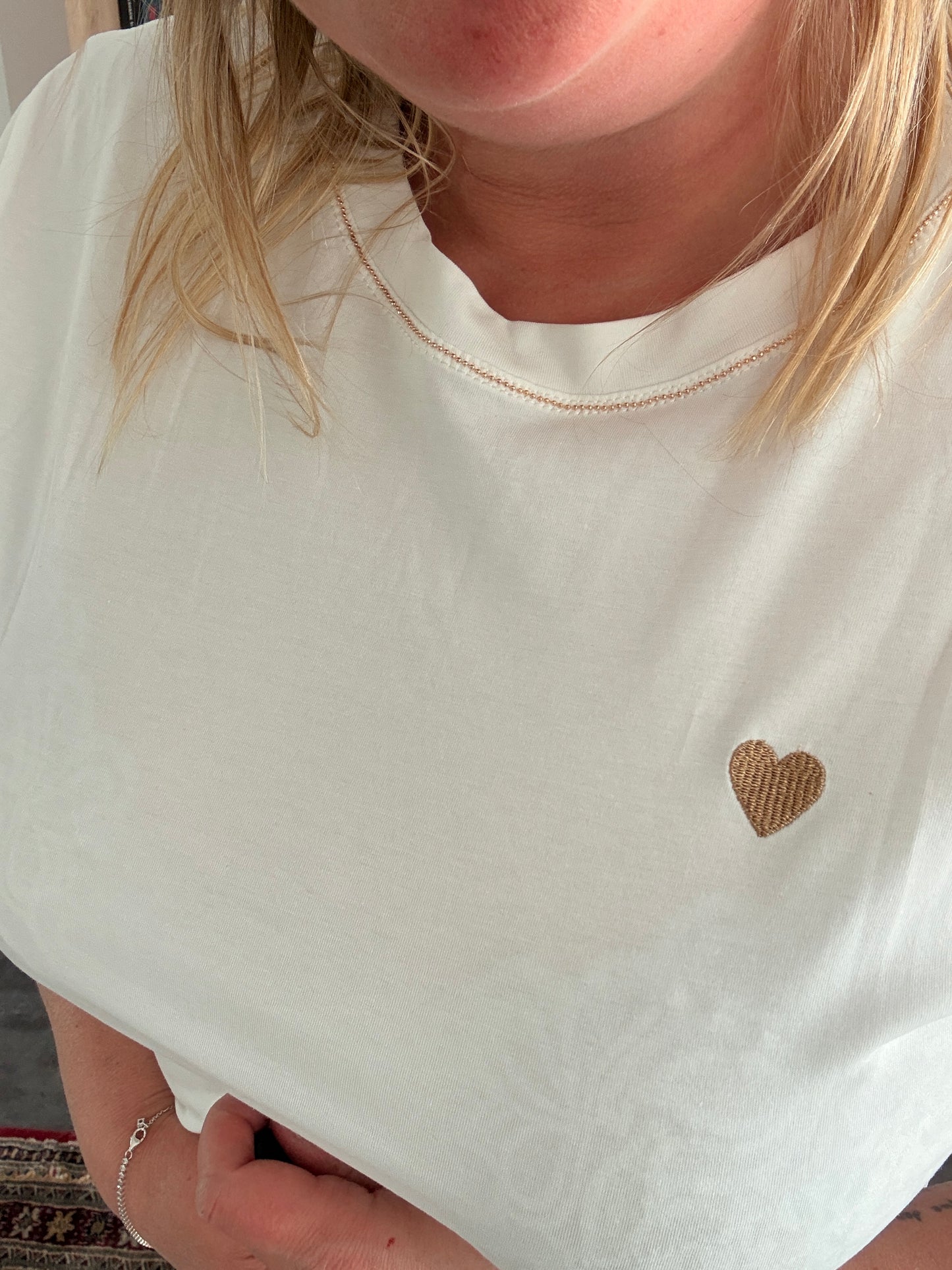 T-shirt blanc avec petit cœur doré sur la poitrine, col rond et détail brillant, femme portant premier plan.