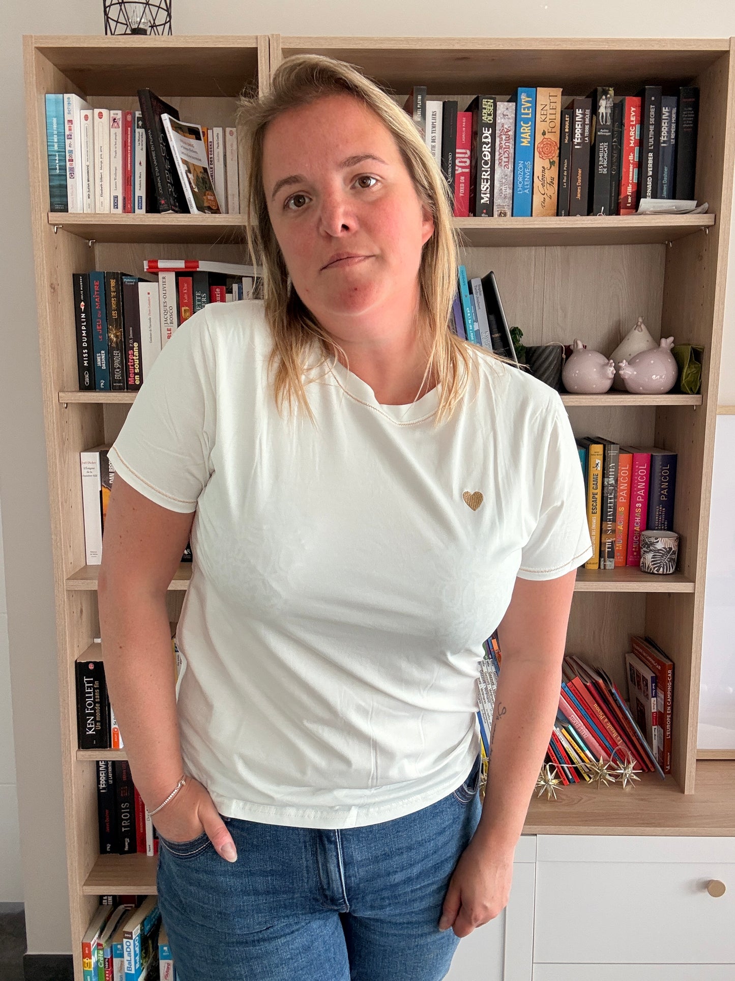 Homme ou femme portant un T-shirt blanc simple avec petit cœur doré sur la poitrine, debout devant une étagère remplie de livres.