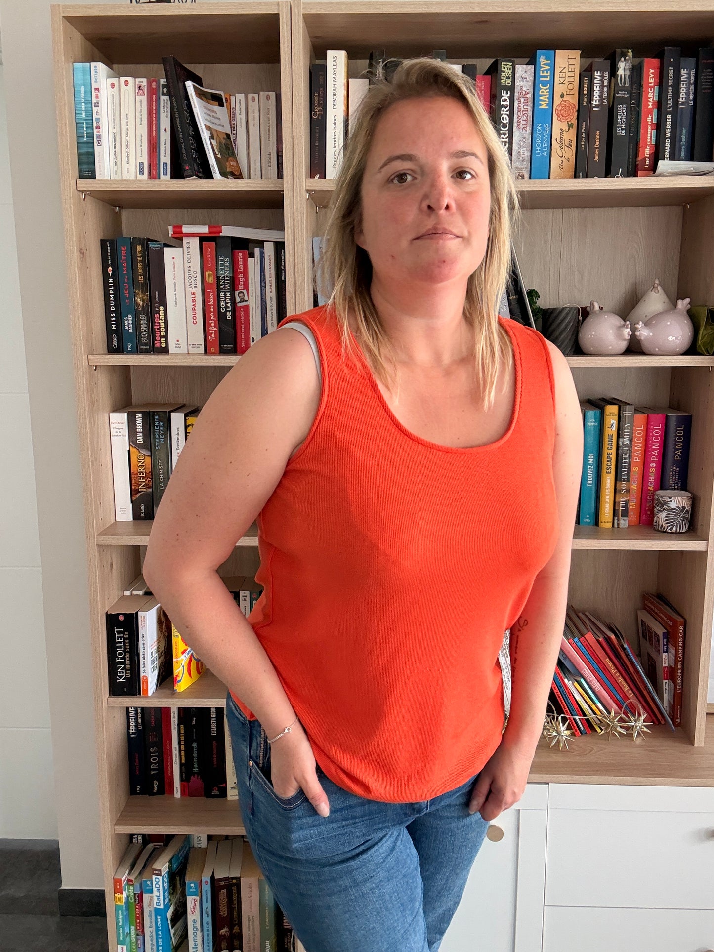 Homme ou femme portant débardeur coton côtelé orange, coupe ample, posé devant une étagère de livres.