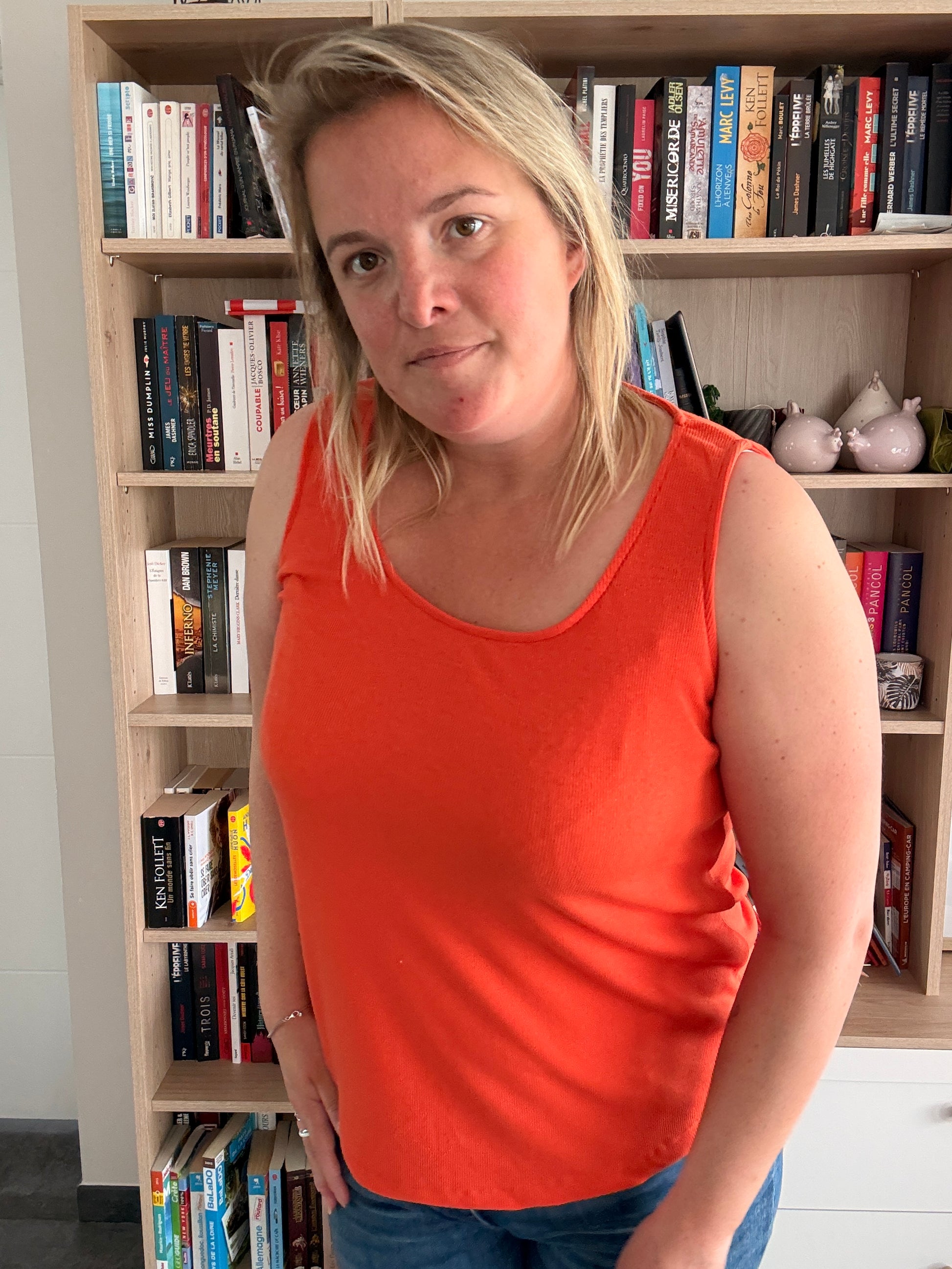 Portait d’une femme portant un débardeur coton côtelé orange, coupe ample et texture douce, debout près d’une bibliothèque.