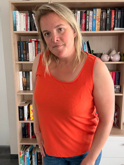 Portait d’une femme portant un débardeur coton côtelé orange, coupe ample et texture douce, debout près d’une bibliothèque.