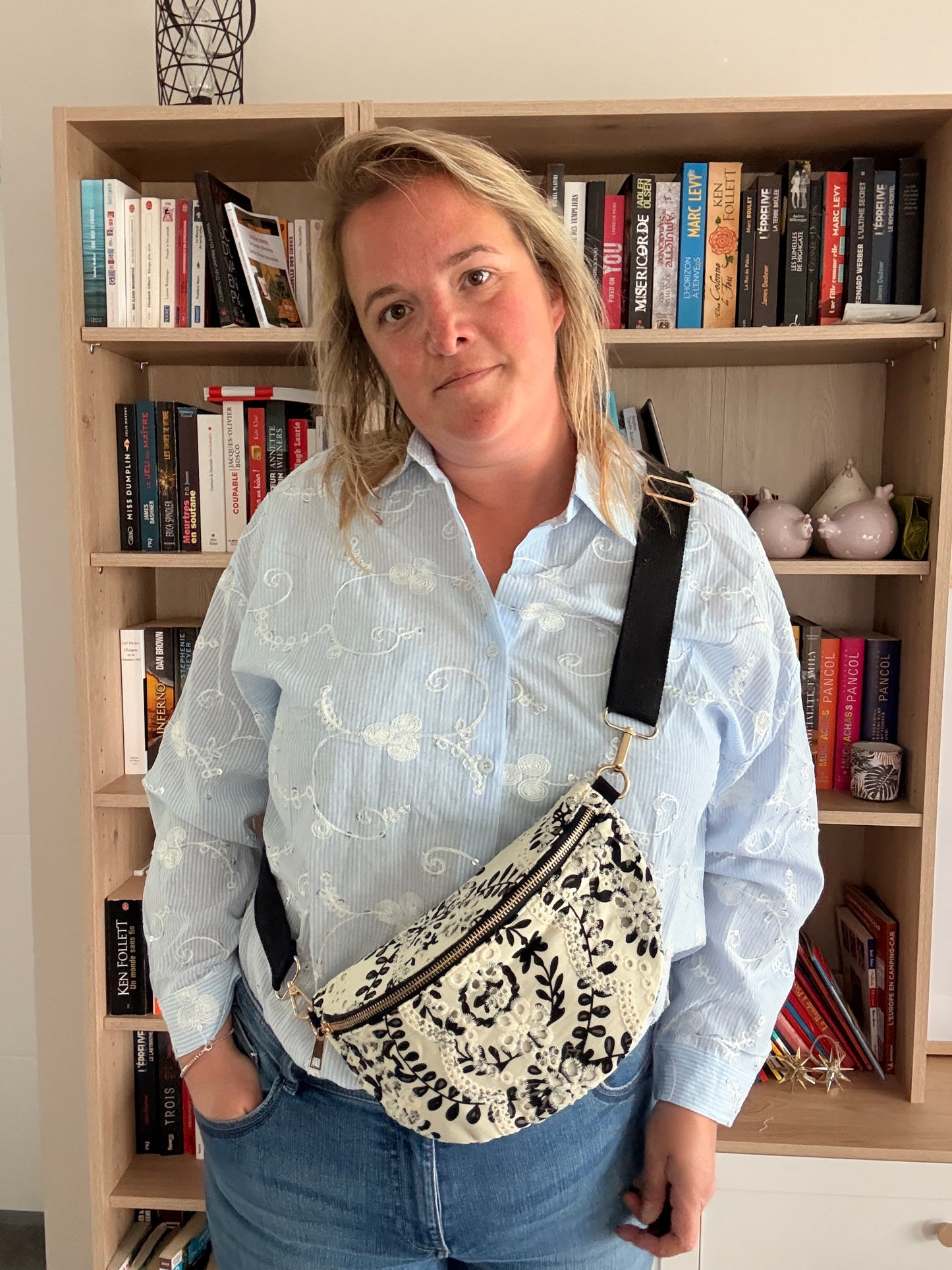 Sac banane femme floral noir et blanc porté en bandoulière, chemise bleu clair et jeans, devant une bibliothèque.