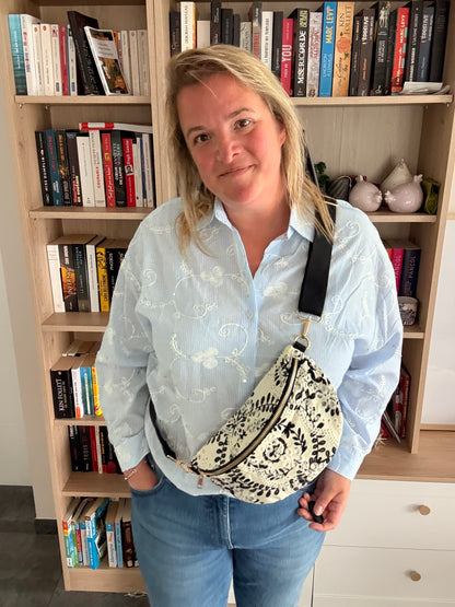 Homme/femme portant sac banane floral noir et blanc légère, stylée et pratique, posé en diagonale sur le torse, devant une bibliothèque.
