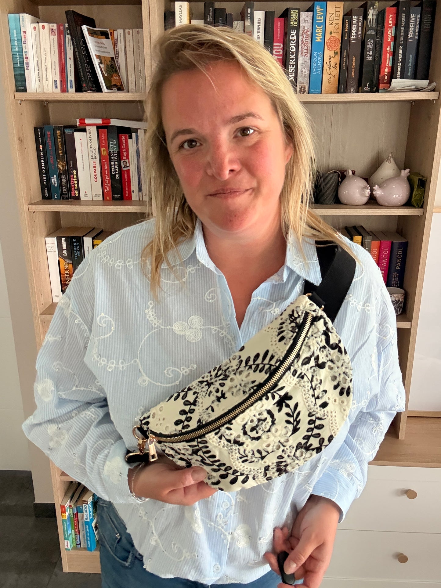 Femme tenant un sac banane floral noir et blanc léger et pratique, porté en ceinture devant une étagère remplie de livres.
