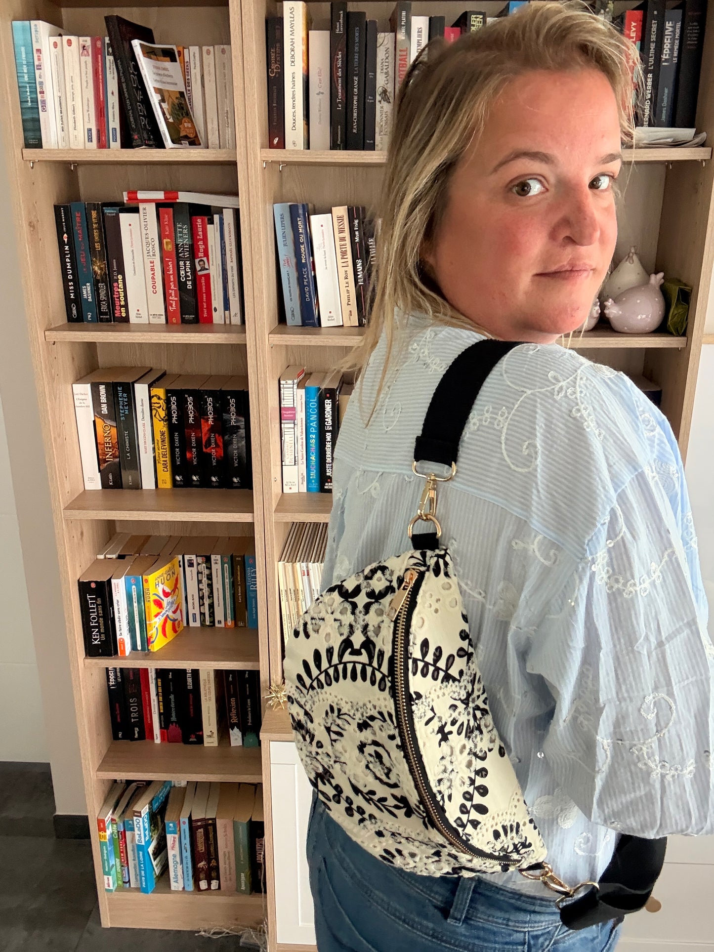 Femme portant un sac banane floral noir et blanc accroché en bandoulière, debout près d'une étagère remplie de livres.