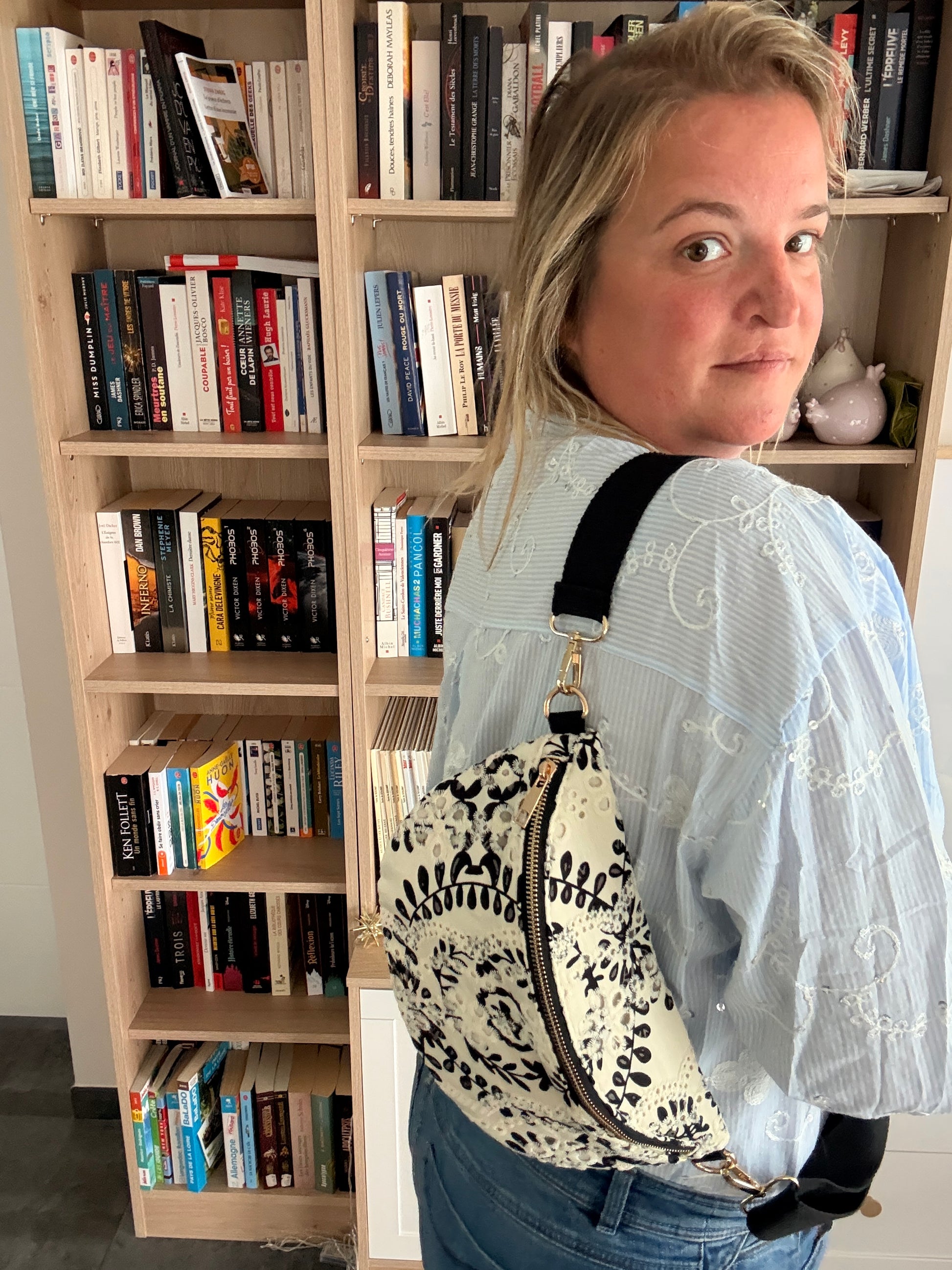 Femme portant un sac banane floral noir et blanc accroché en bandoulière, debout près d'une étagère remplie de livres.