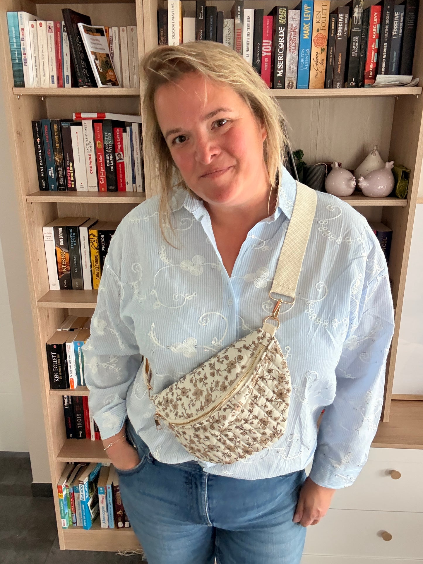 Sac banane femme fleurie taupe avec fond ivoire porté en bandoulière réglable, doré zip et look casual devant étagère de livres.